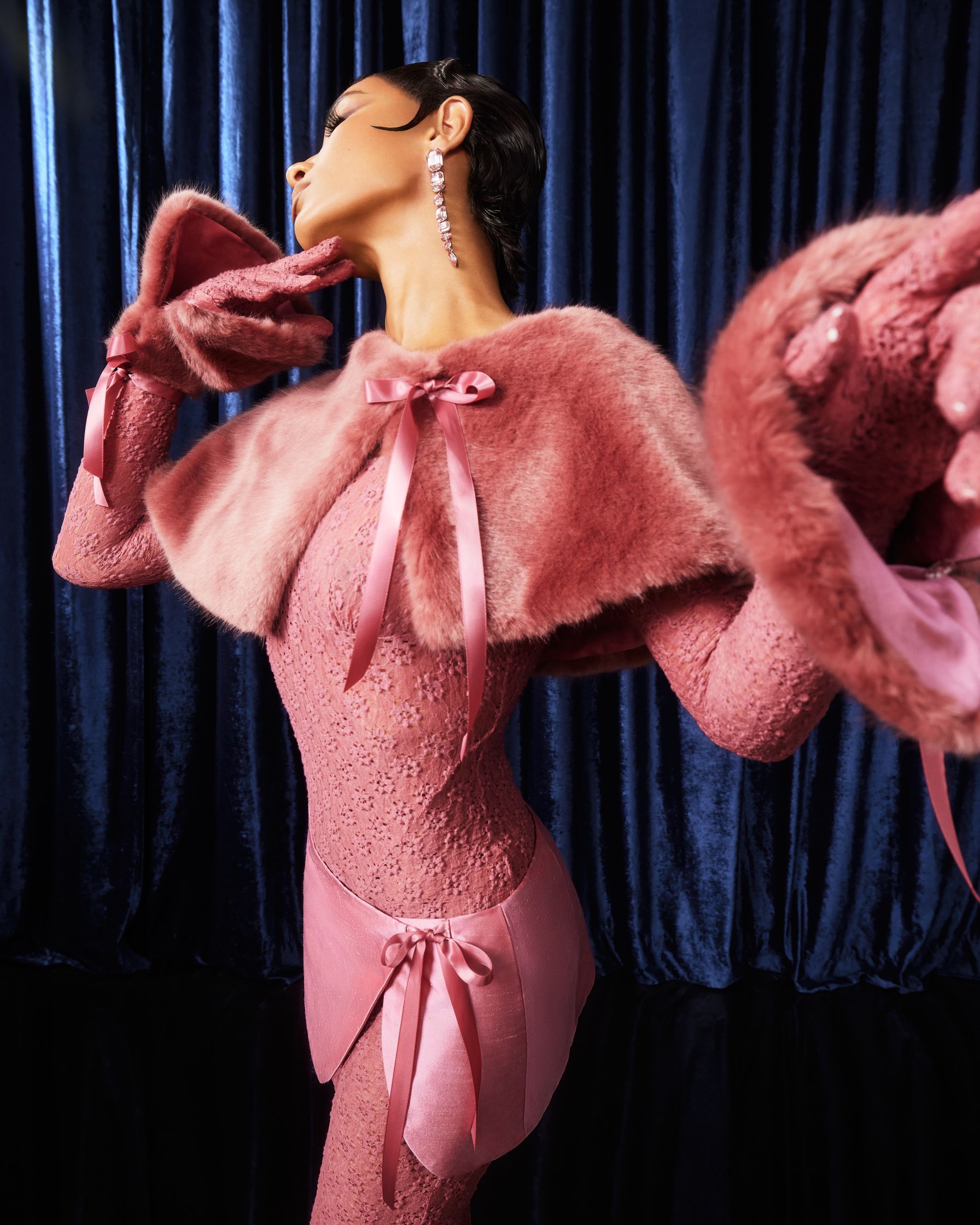 a woman in a pink fur coat posing in front of a curtain