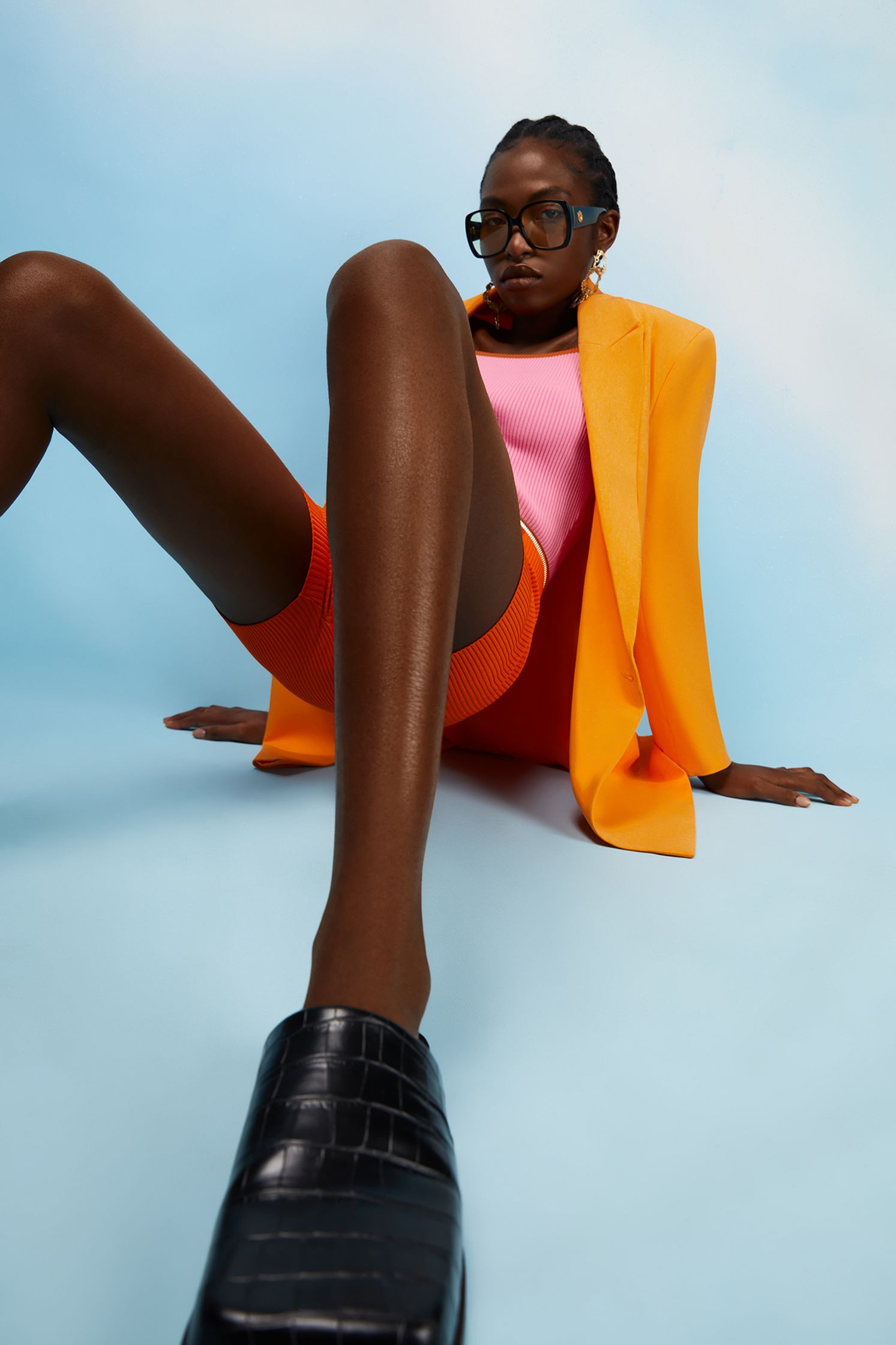 a black woman in an orange blazer and shorts laying on a blue background