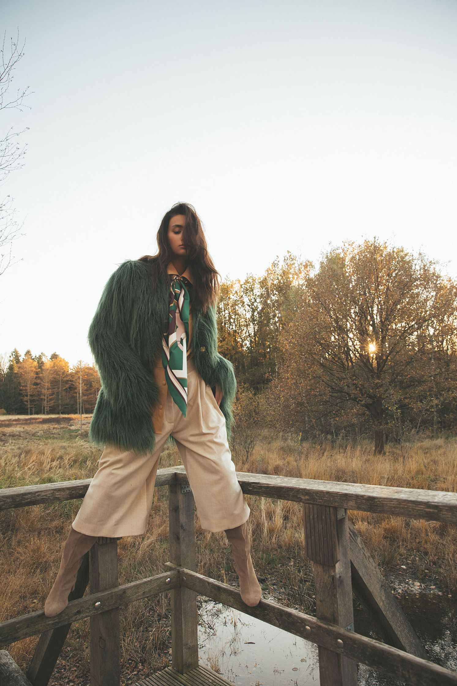 a woman in a green fur coat standing on a wooden bridge
