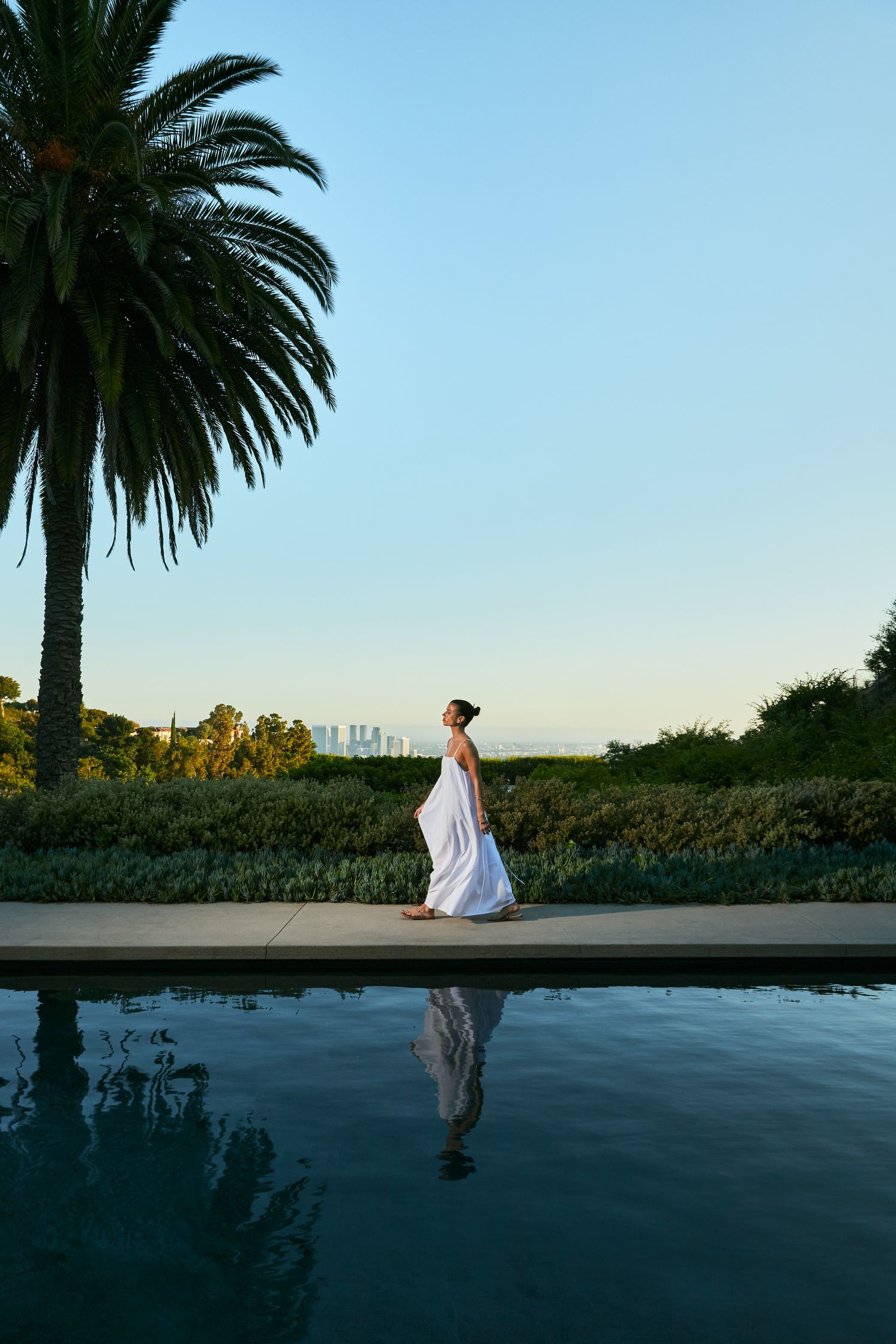 a woman in a white dress walking by a pool with palm trees in the background