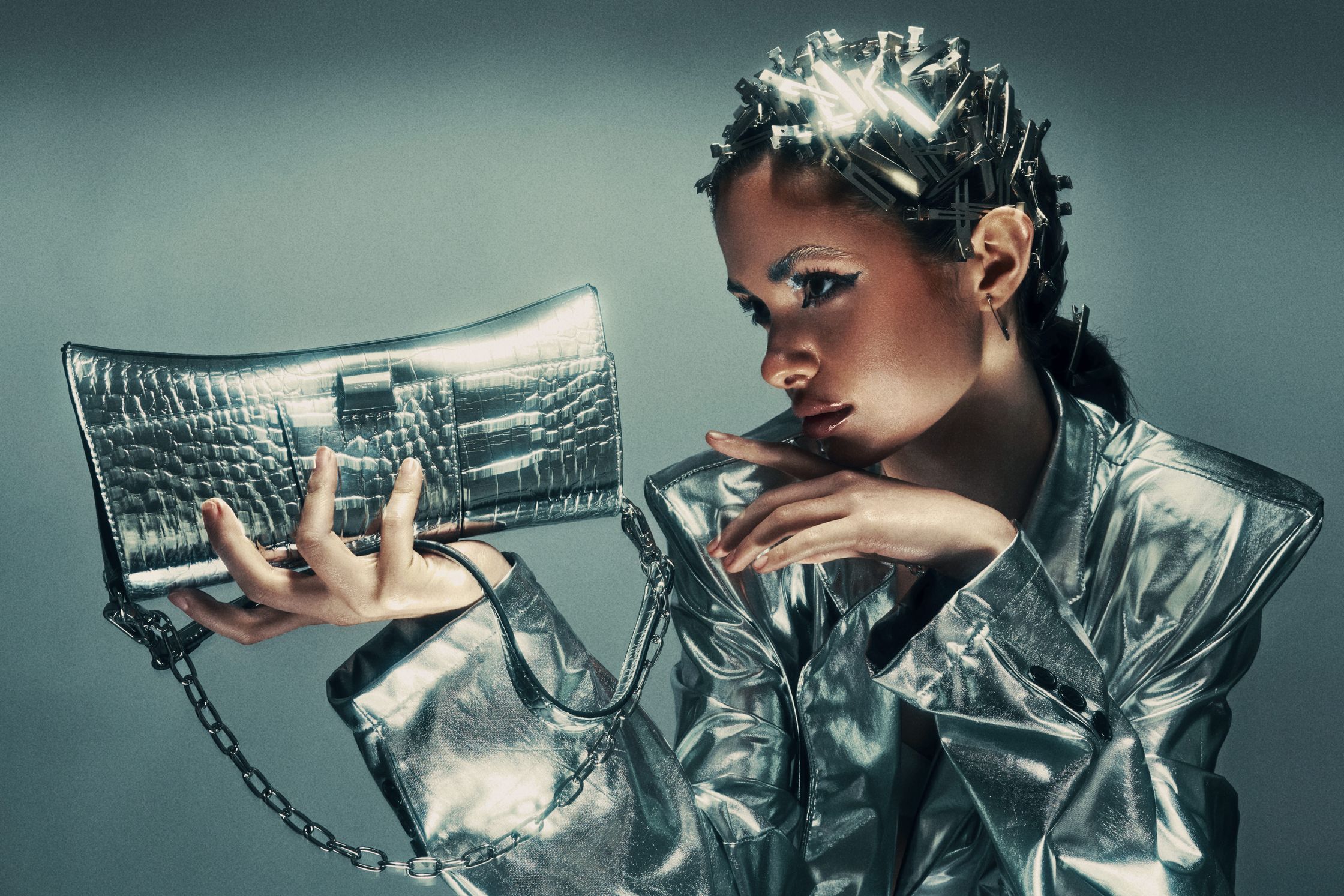 Dramatic close-up of Monique Bourscheid with bold makeup and silver hair accessories, resting her chin on her hand while holding a metallic handbag 