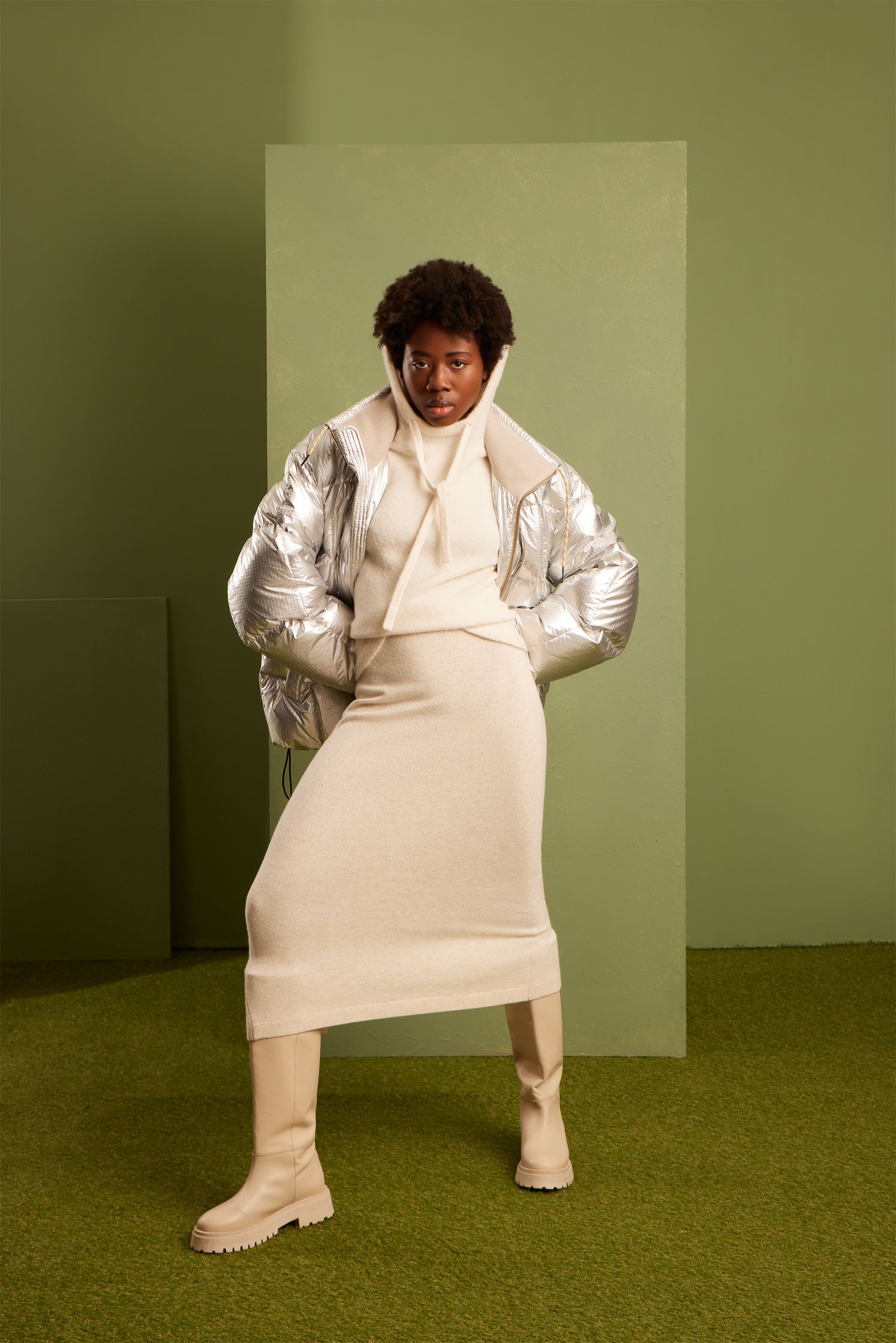 a woman in a white jacket and boots posing in front of a green wall