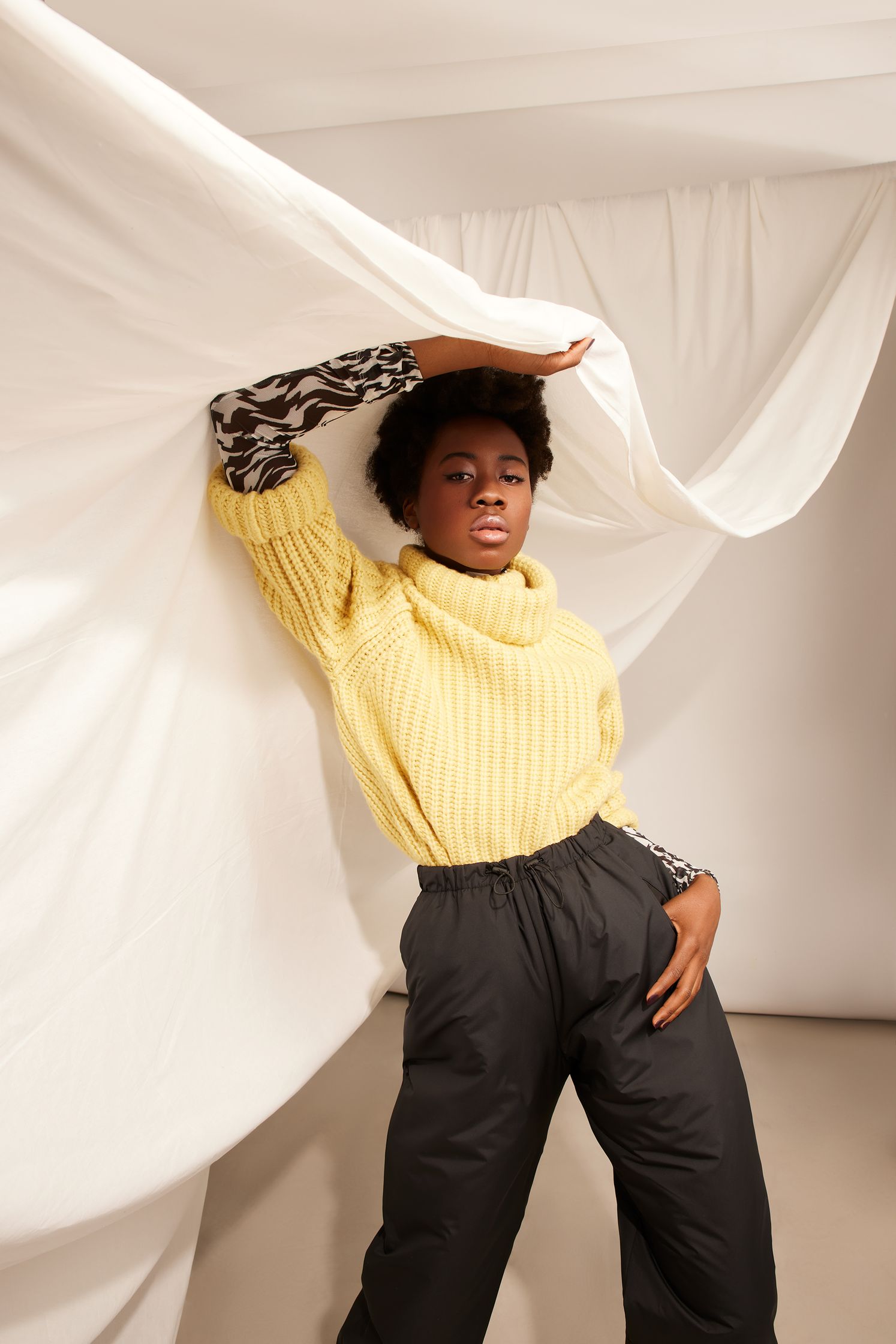 a black woman in a yellow sweater posing in front of a white sheet
