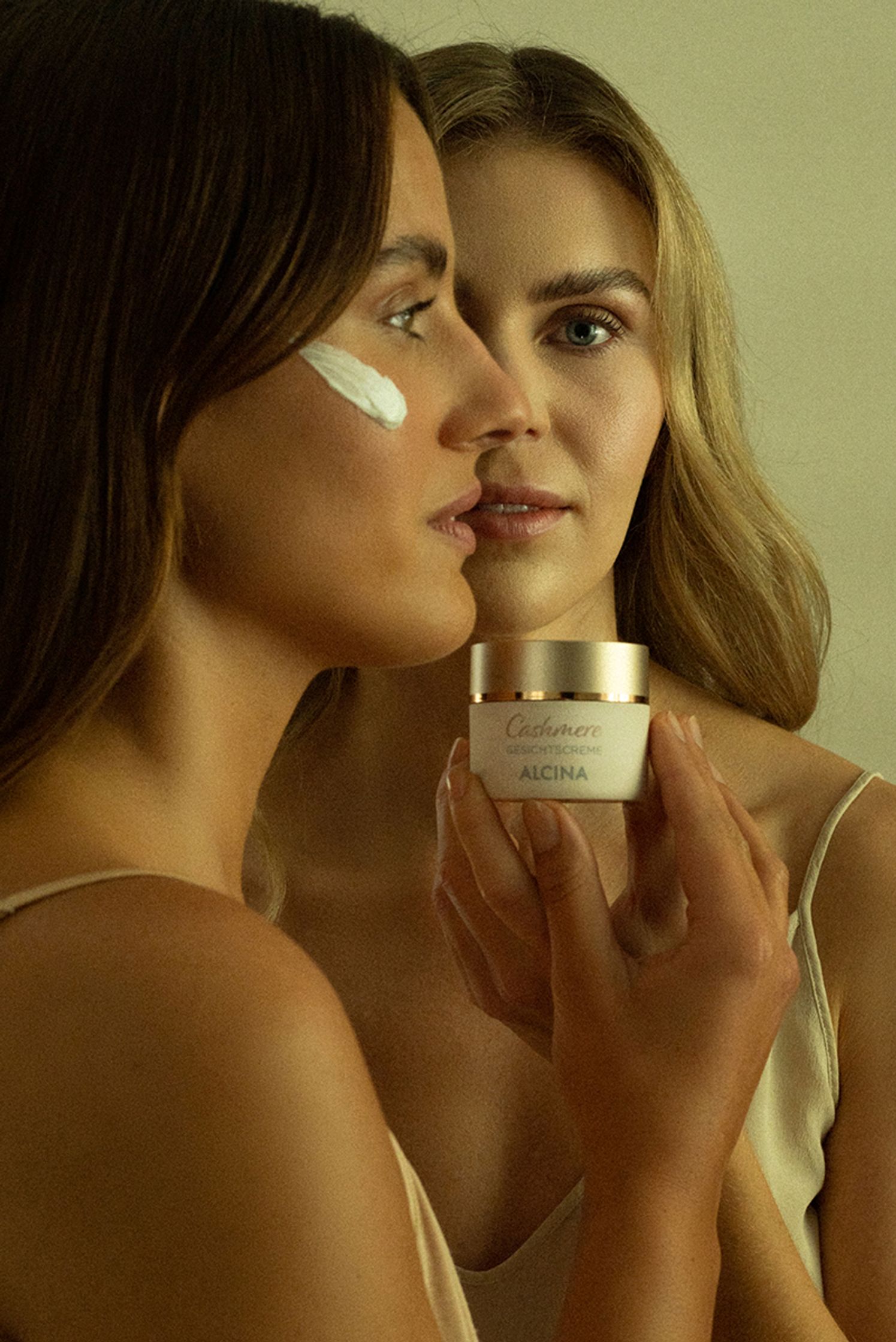two women looking at a alcina cream jar in front of a mirror