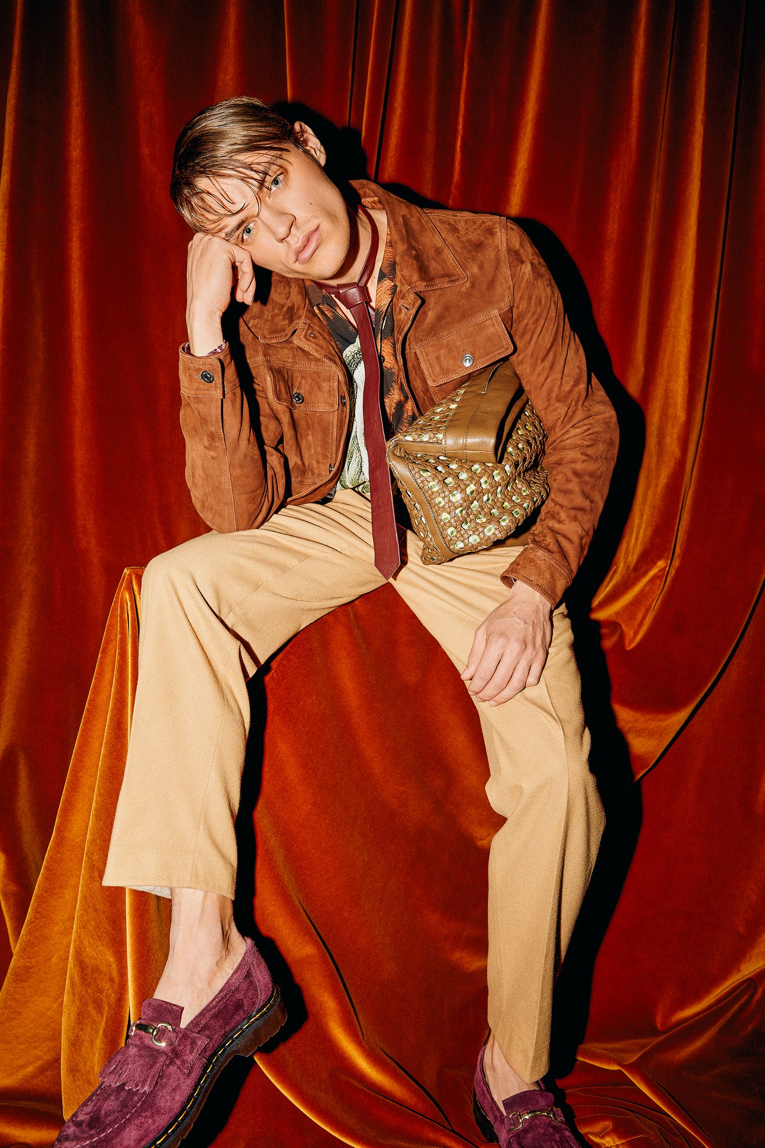 a man in a brown jacket and brown pants sitting on a red curtain