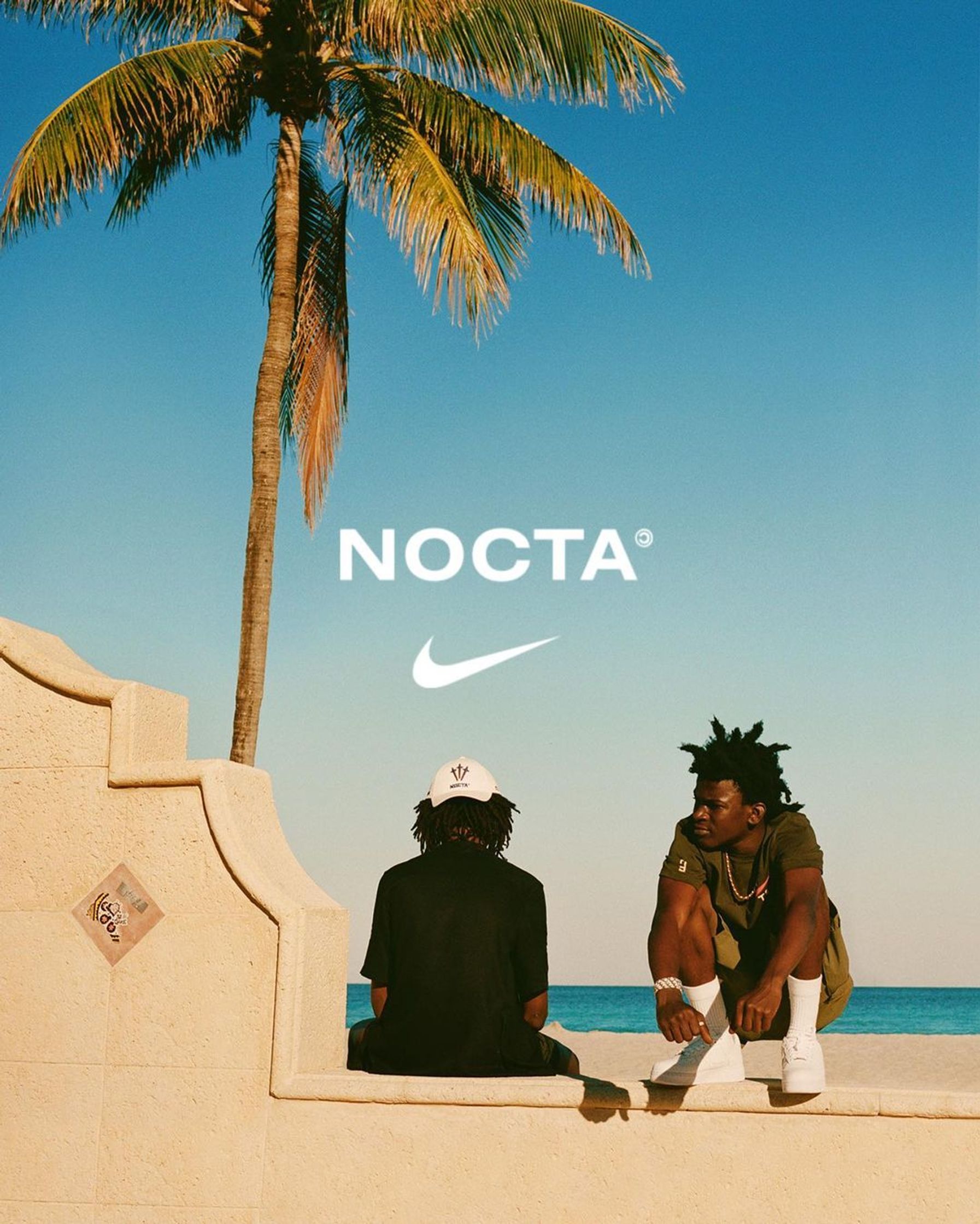 two men sitting on a wall next to a palm tree nike