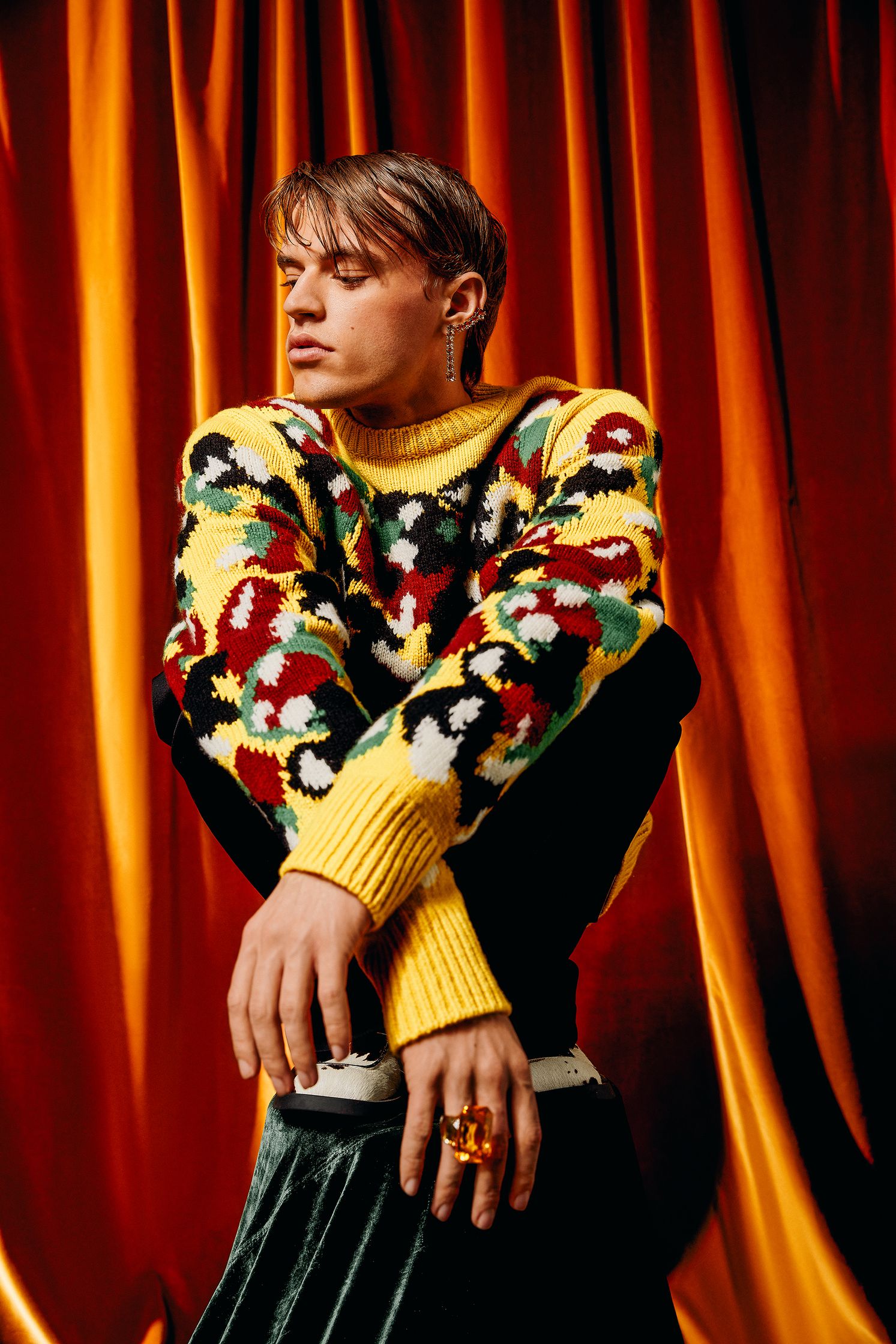 a young man in a colorful sweater sitting on top of a red curtain