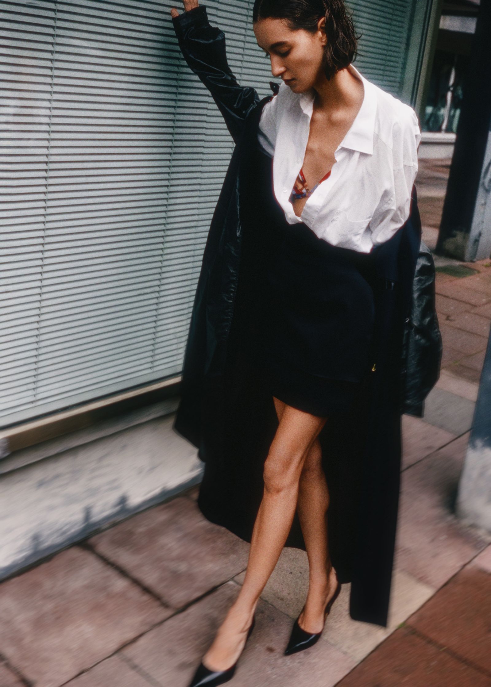 a woman in a white shirt and black coat leaning against a wall