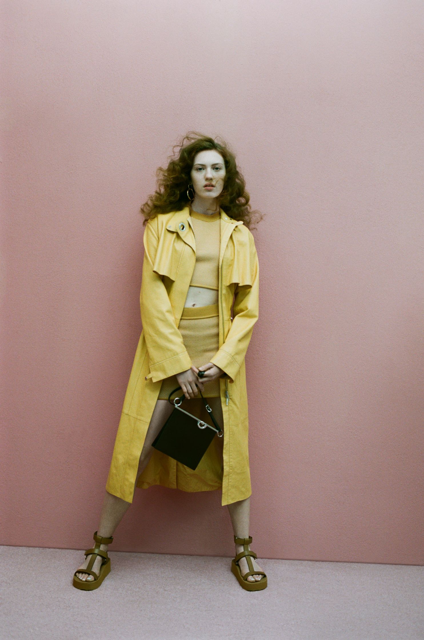 a woman in a yellow coat posing against a pink wall