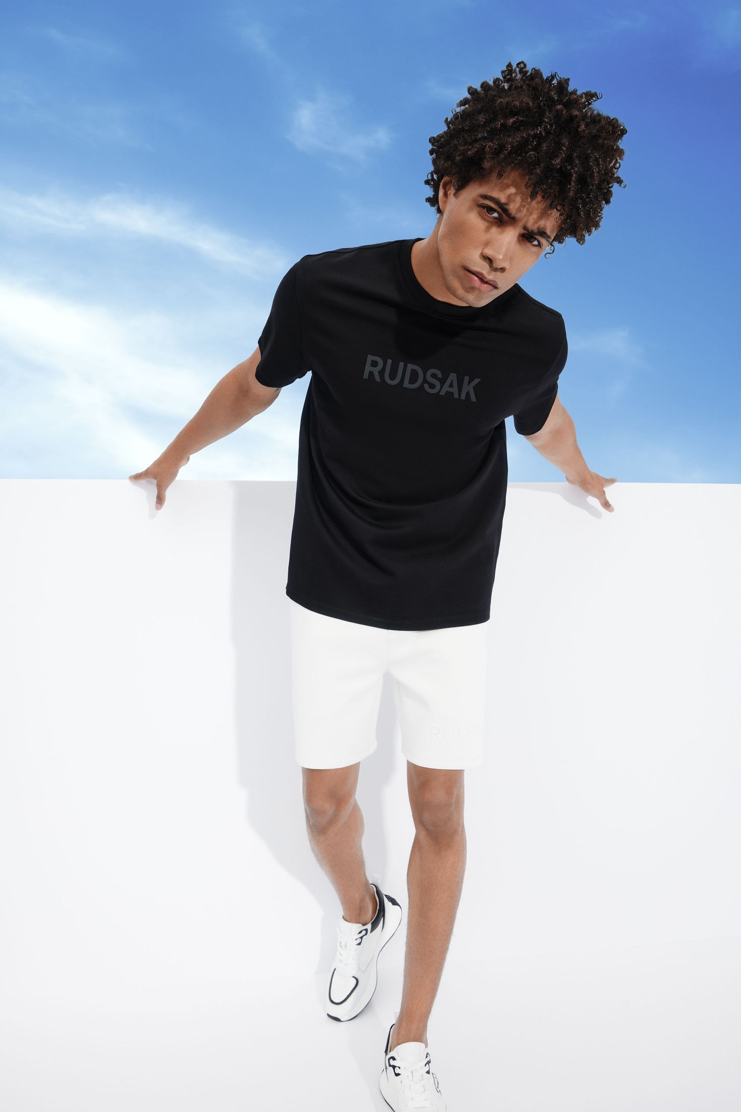 a man in a black t - shirt and white shorts leaning against a wall