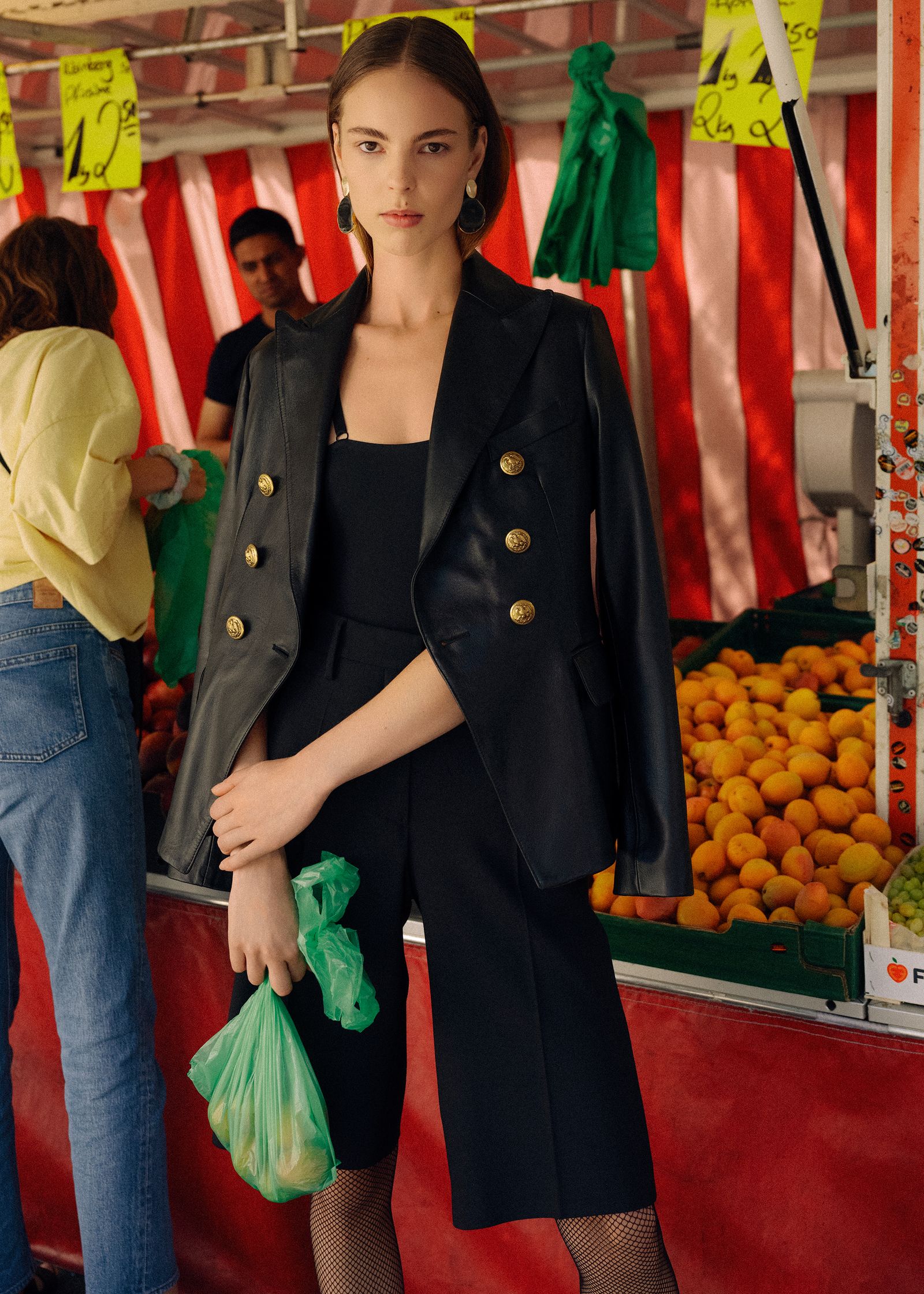 a woman in a black blazer standing in front of a fruit stand