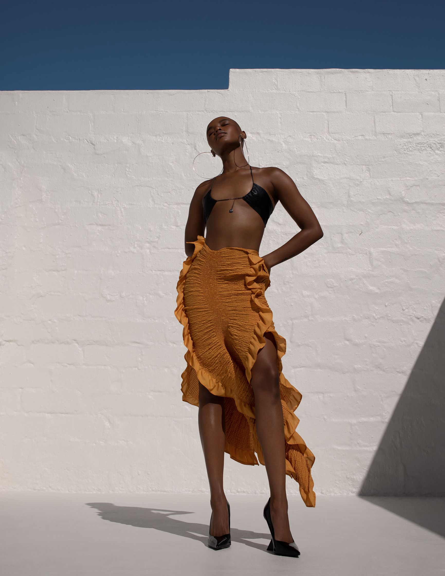 a black woman in a bikini and ruffled skirt posing in front of a white wall