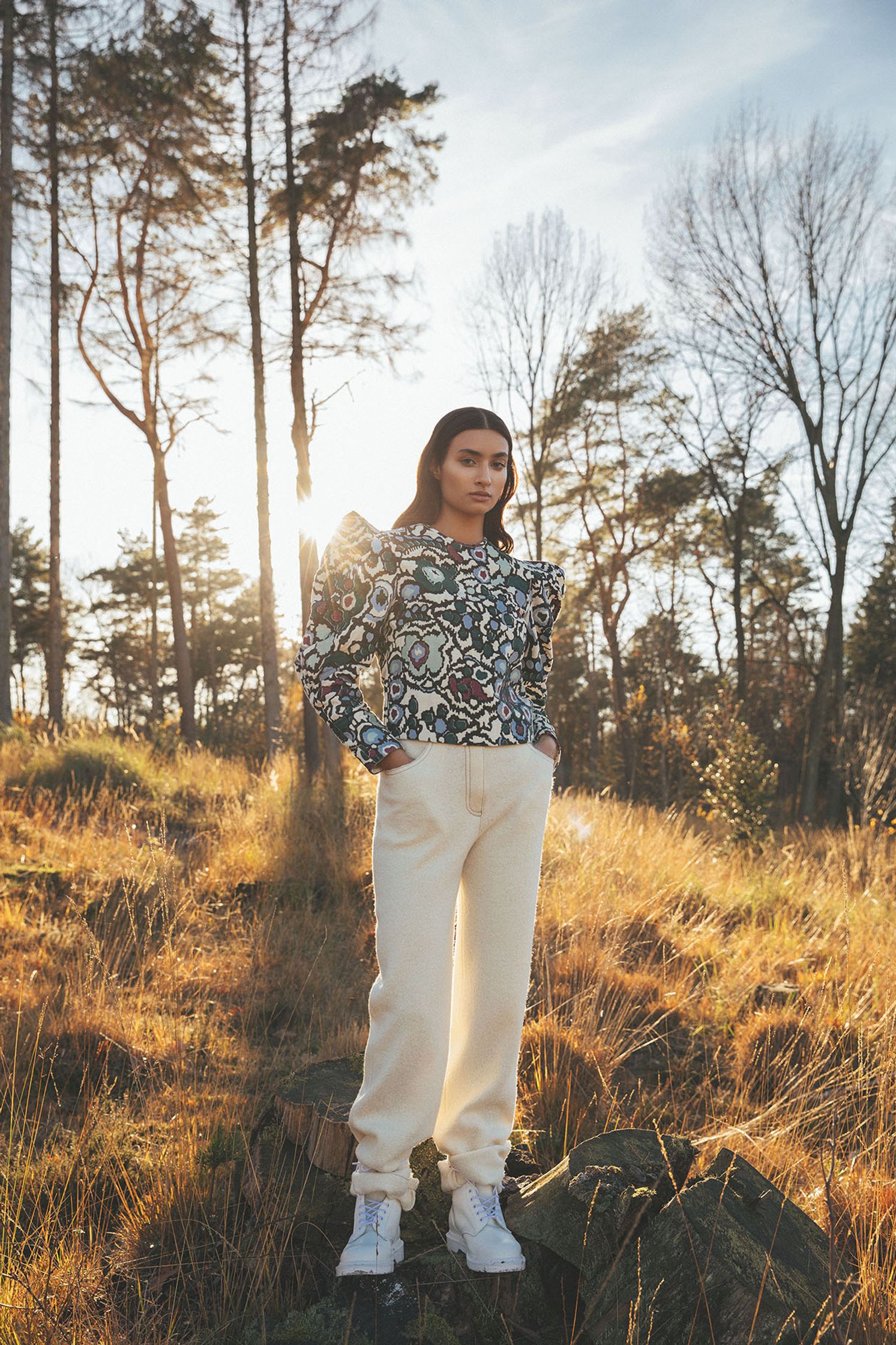 a woman standing in the woods wearing white pants and a floral top