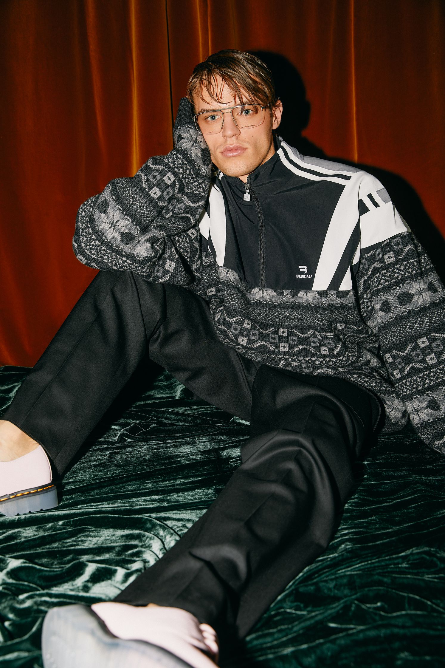 a man sitting on a bed wearing a sweater and sneakers