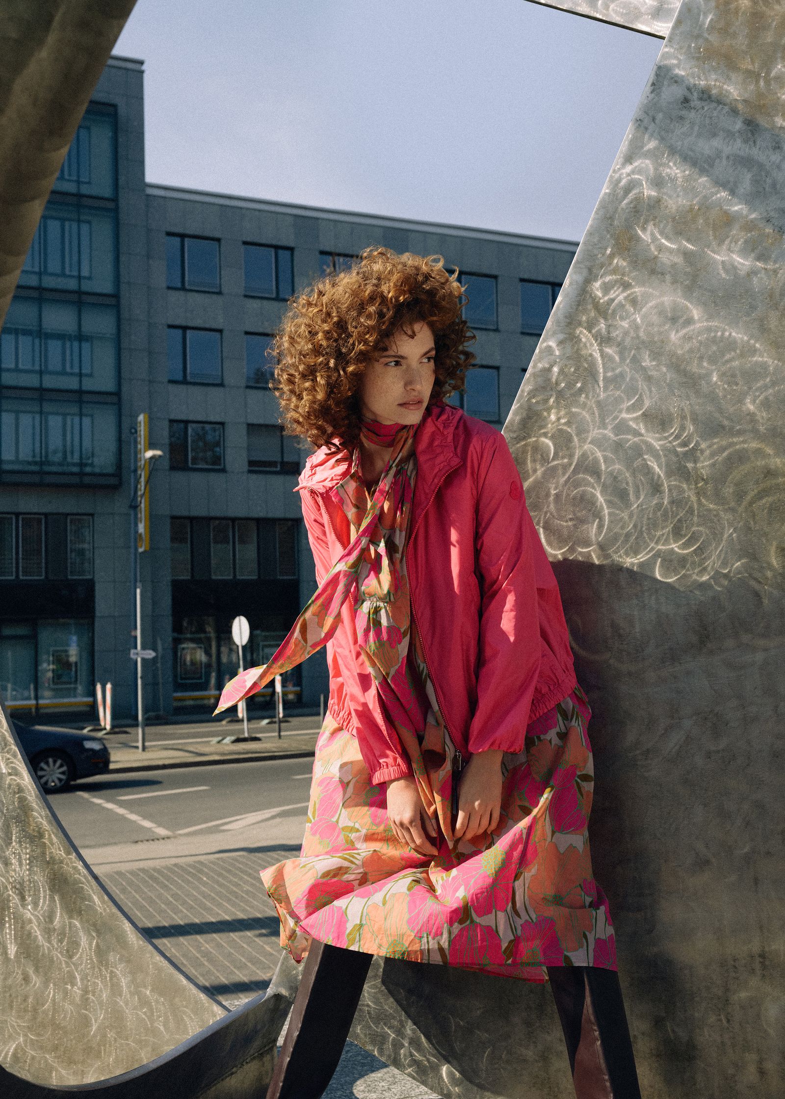 a woman in a pink jacket leaning against a metal sculpture