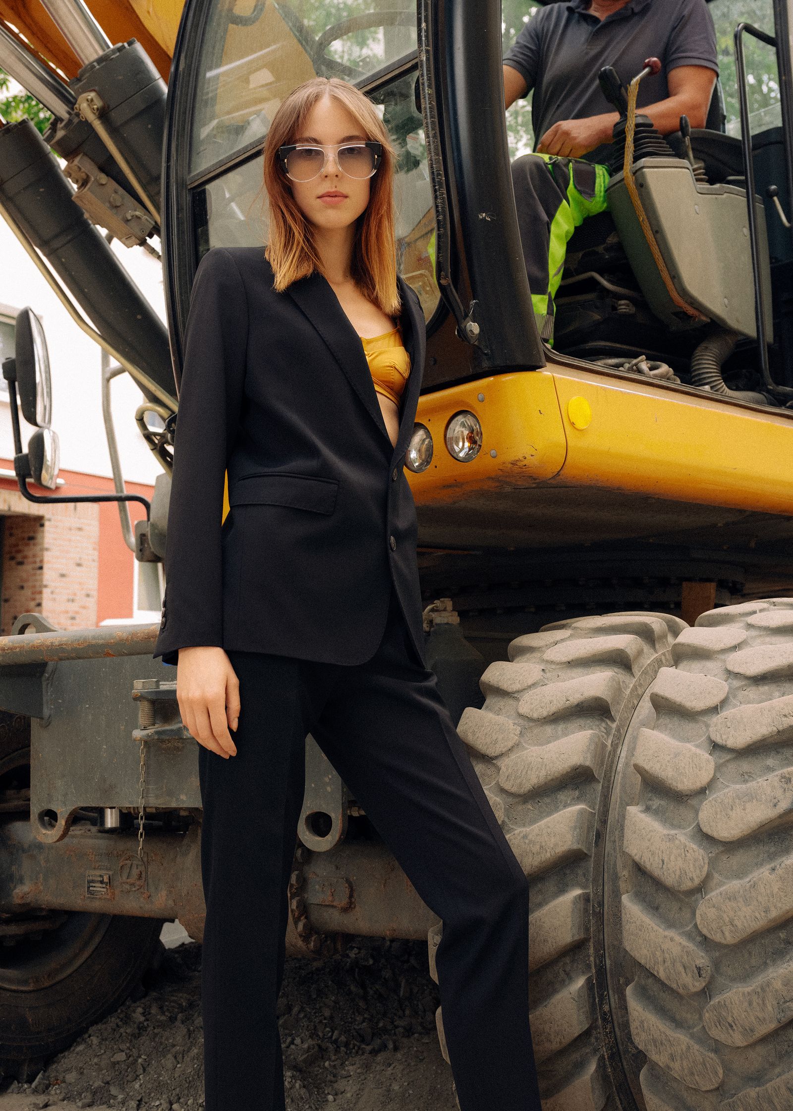 a woman in a black suit standing next to a yellow excavator