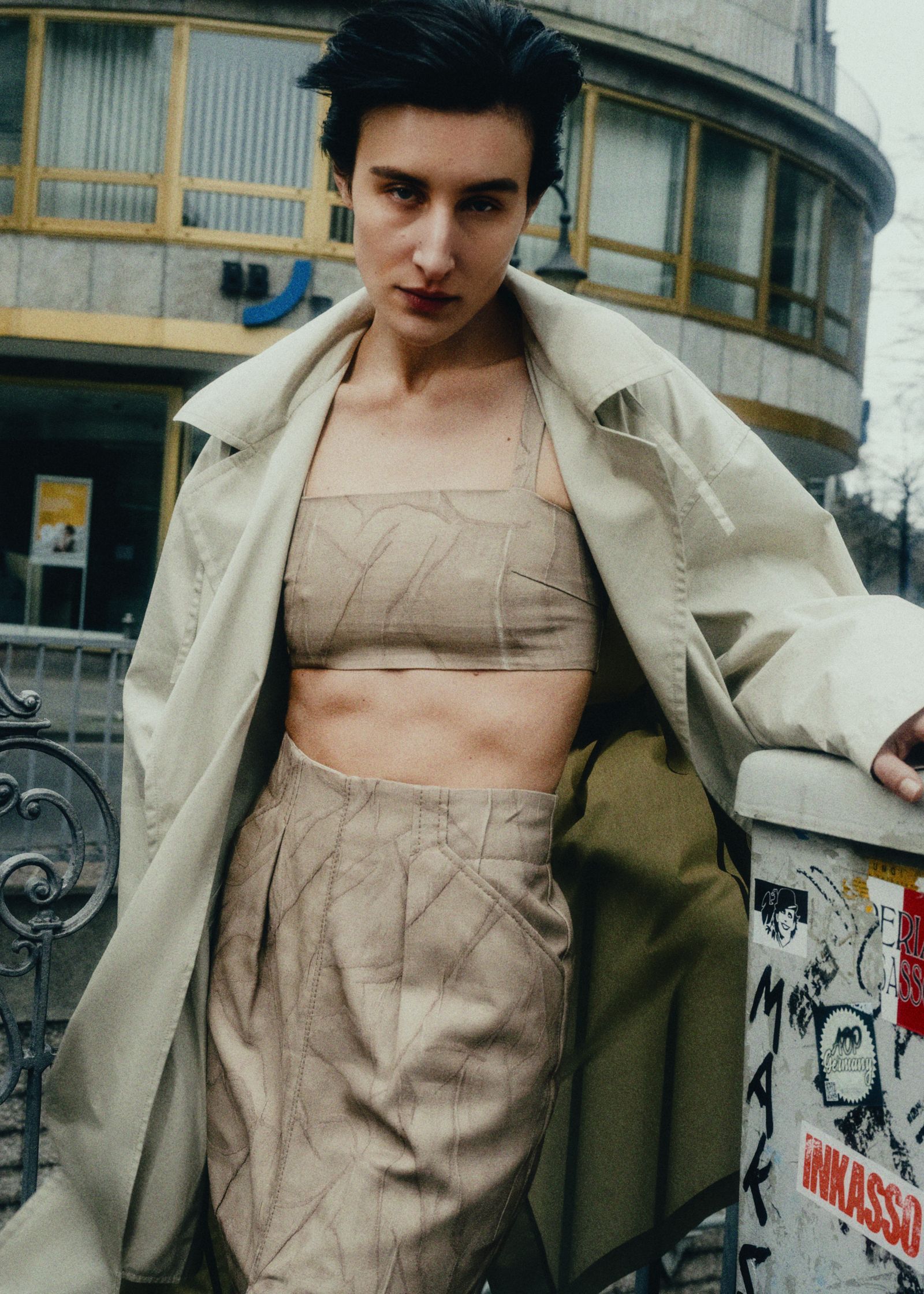 a woman in a tan coat and skirt leaning against a wall