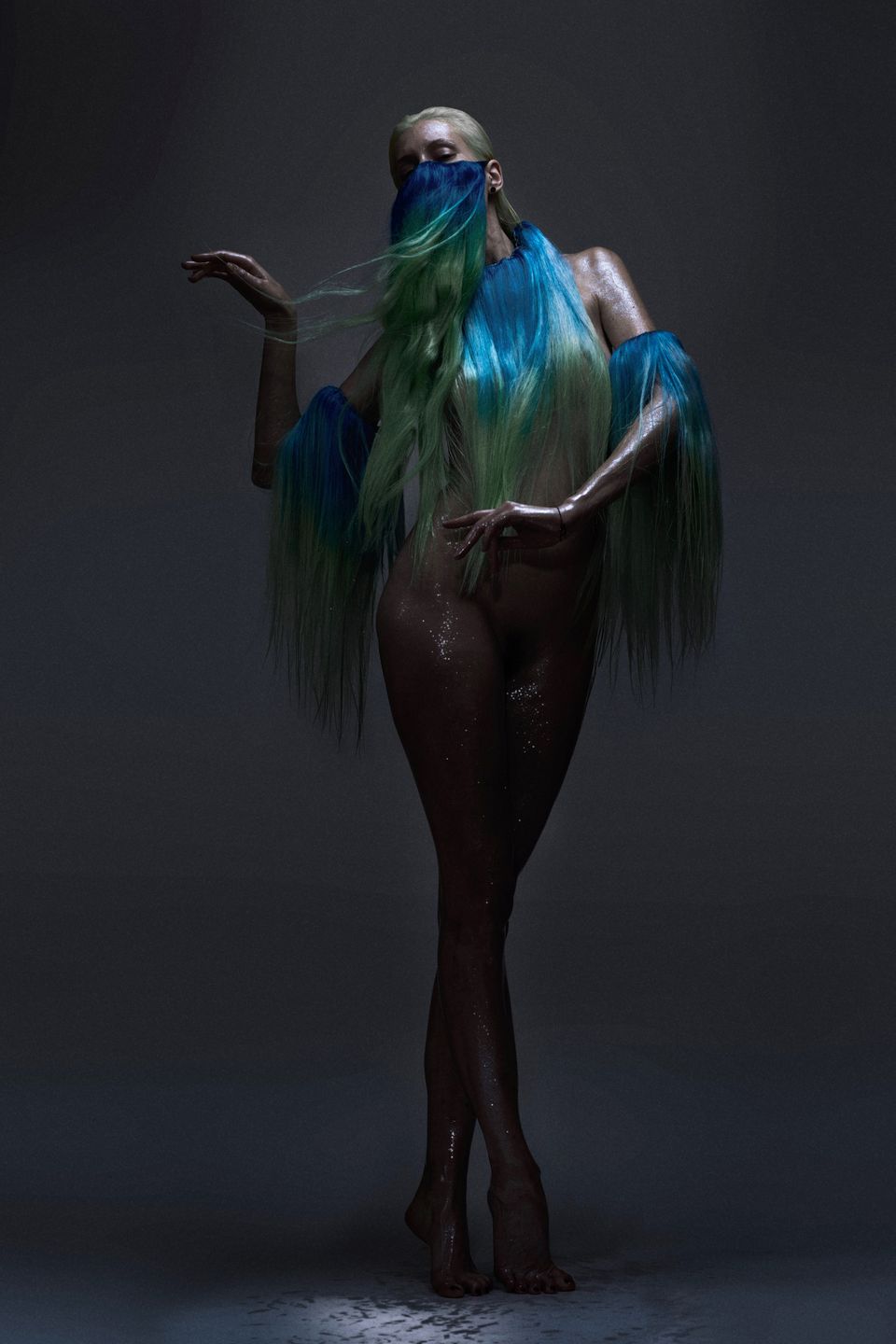 Editorial shot of a model wearing a hairpiece as clothing, showcasing vibrant blue and green tones against a dark, moody background.
