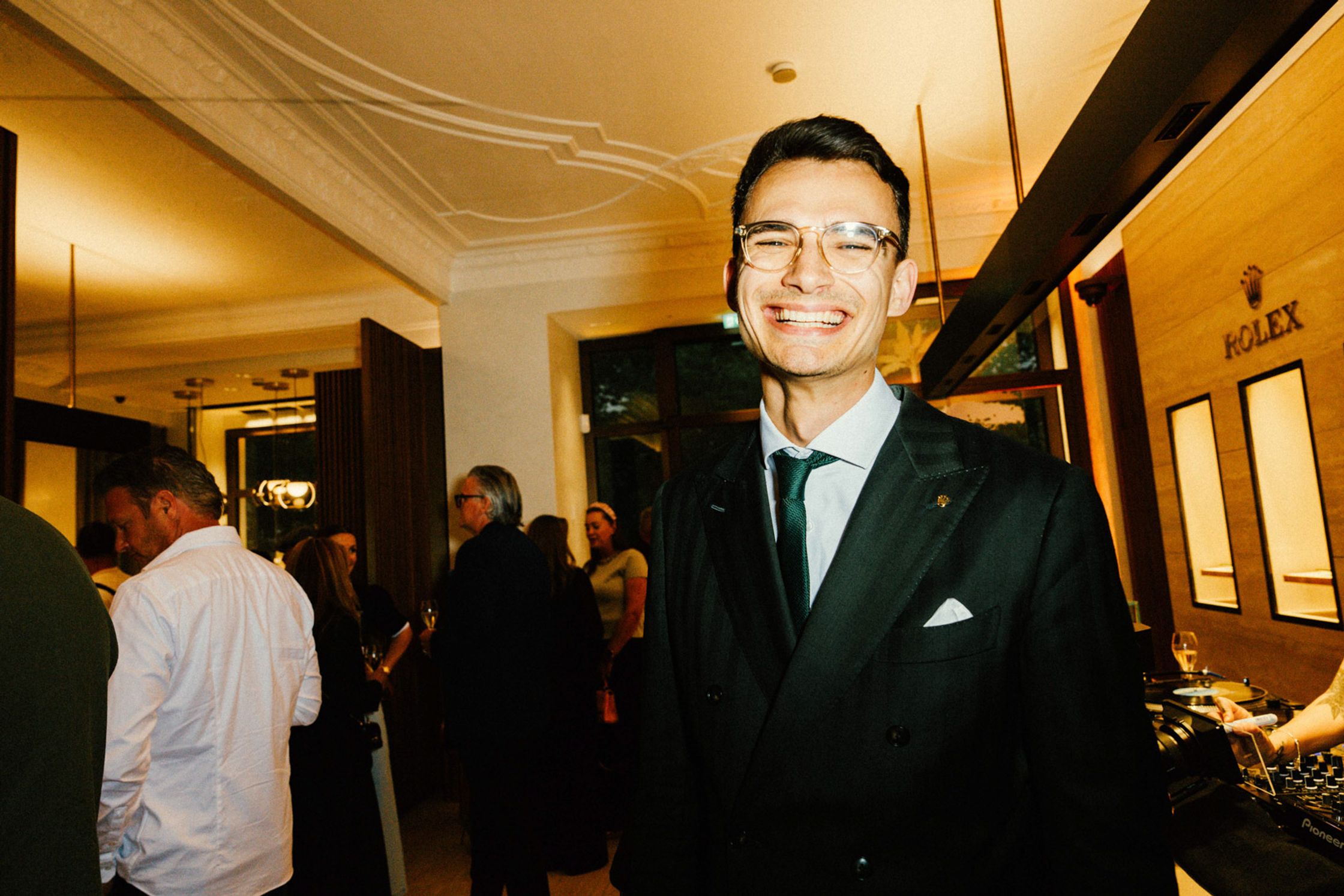 Eleganter Mann im dunklen Anzug mit grüner Krawatte lächelt strahlend beim exklusiven Rolex Boutique Event Berlin, Pioneer DJ-Pult und Rolex Signage im Hintergrund