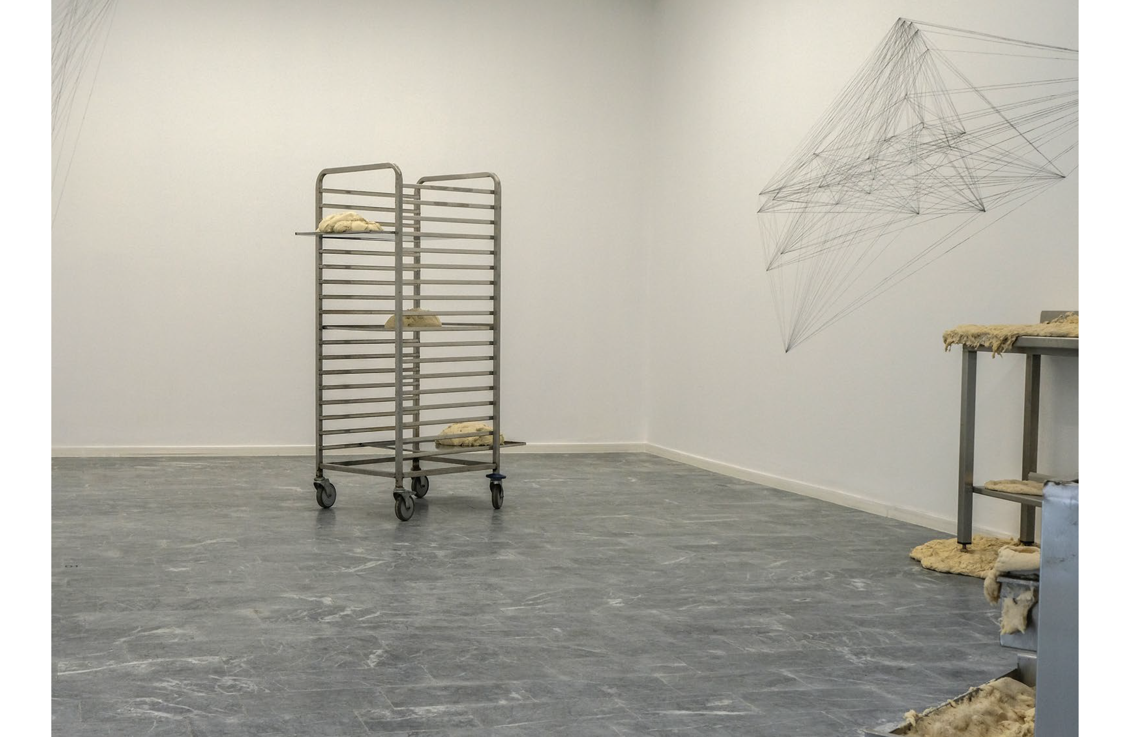 Metal baking racks filled with dough sit next to an abstract string art installation in a minimalist gallery.