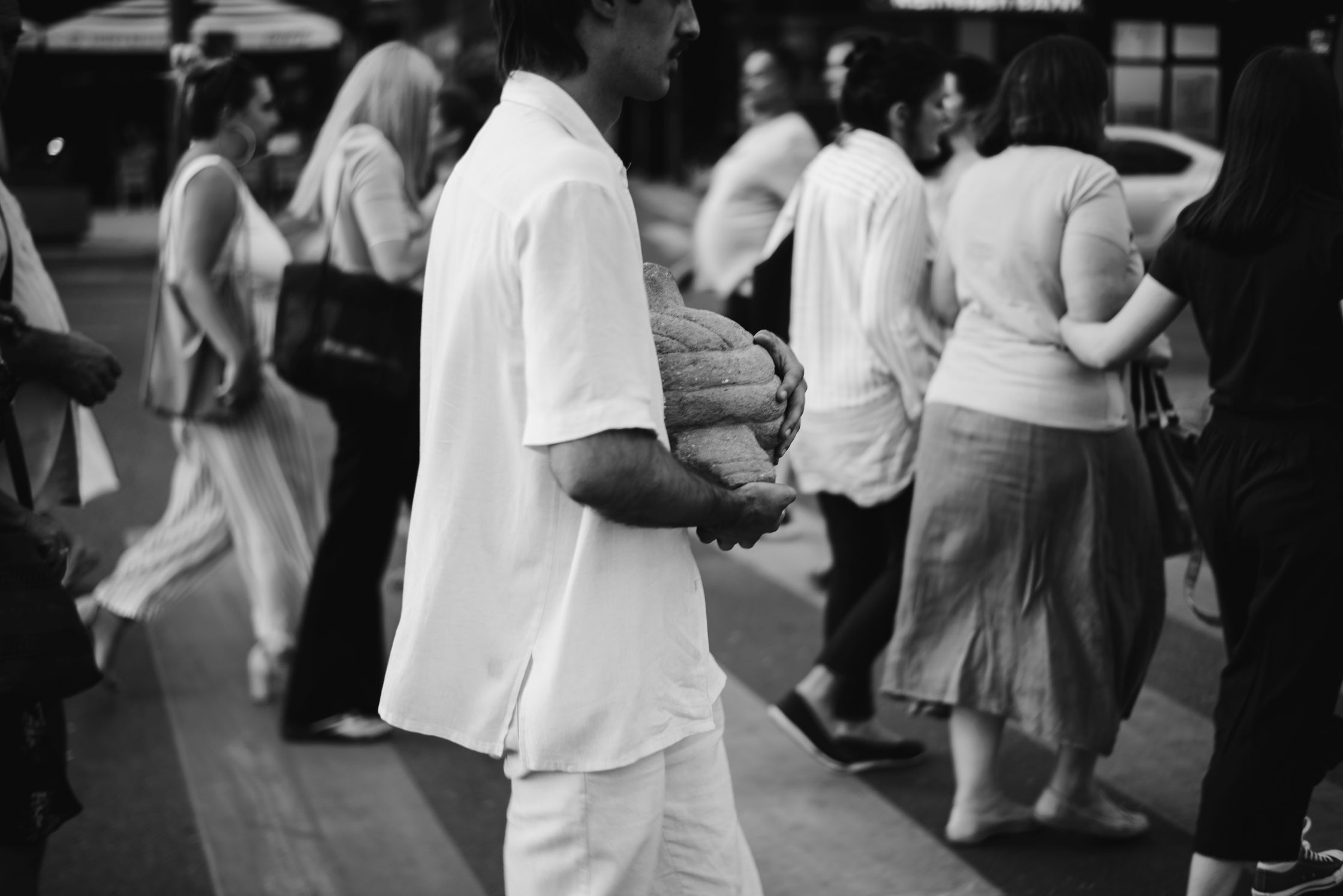 A grayscale, eye-level photo shows people crossing a street, with a man carrying a small pumpkin or melon in the foreground.