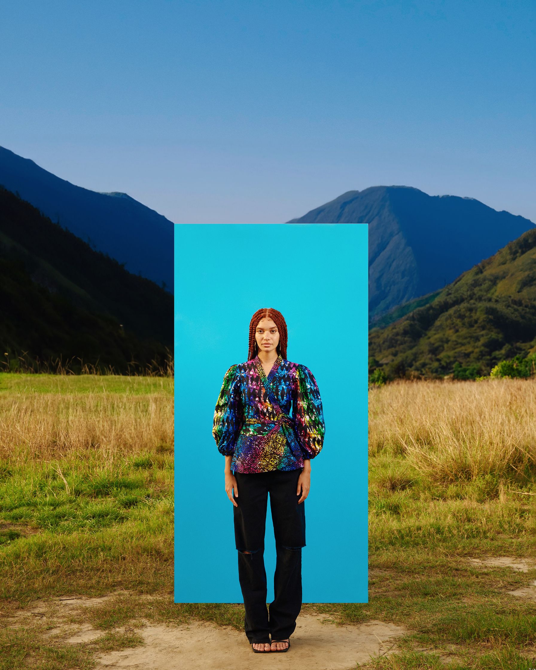a woman standing in front of a blue board in a field with mountain behind wearing an african attire photographed by Faith Adu Afriyie