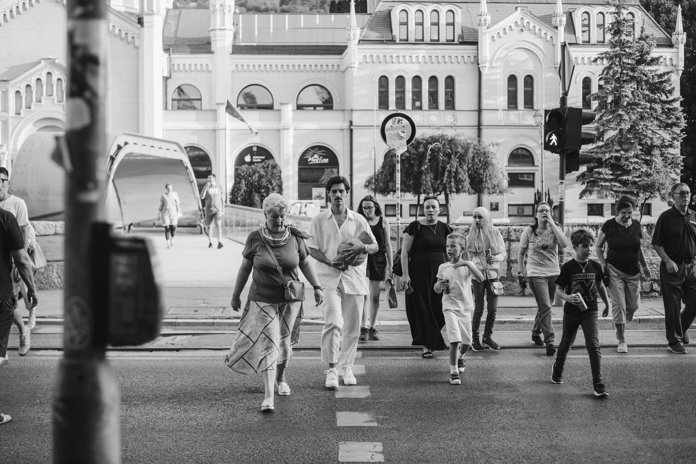 A black and white street photograph of people walking across a street with a building and trees in the background.