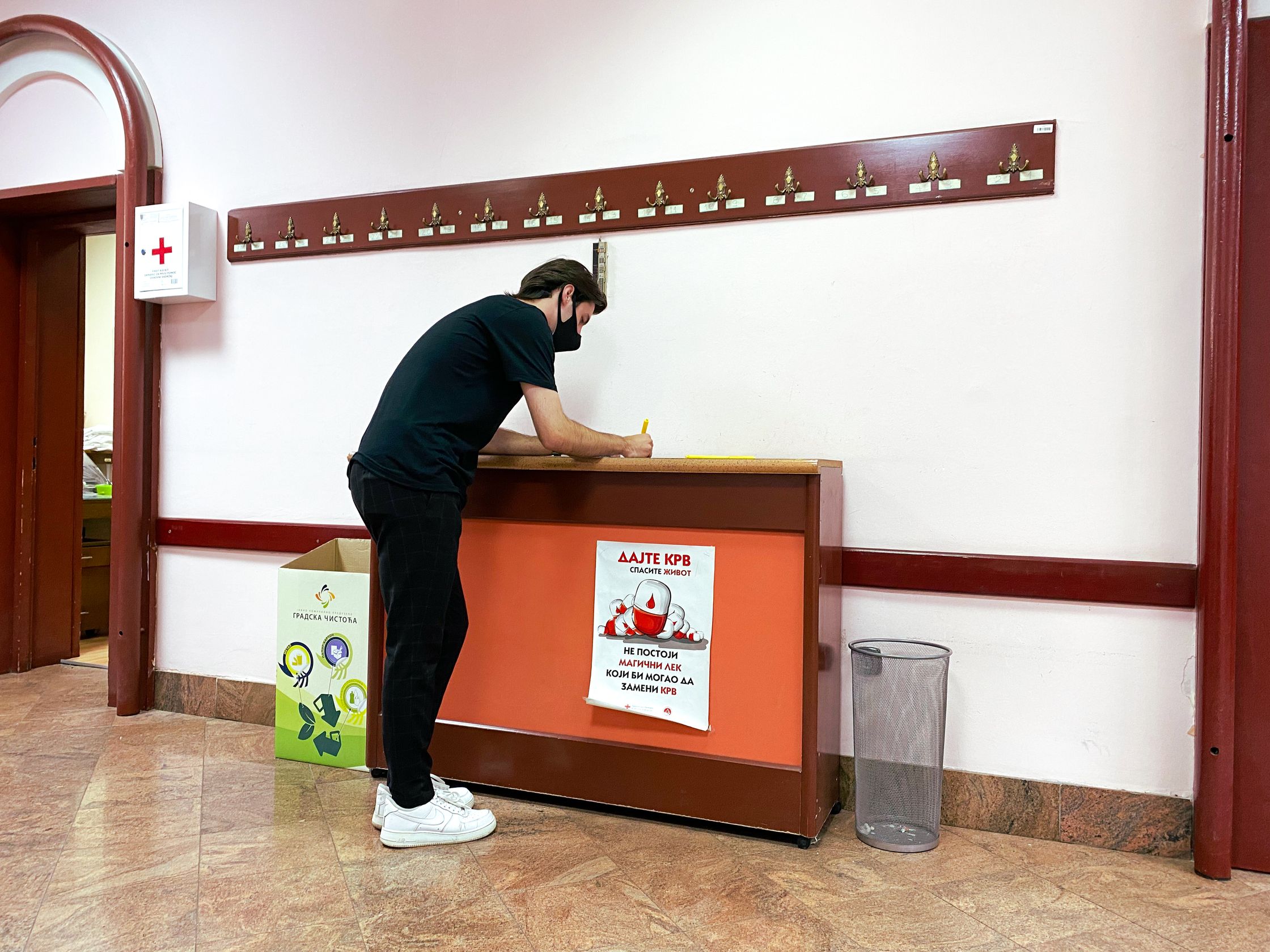 A masked person leaning over a counter in a room with a poster that reads "Give Blood, Save a Life."