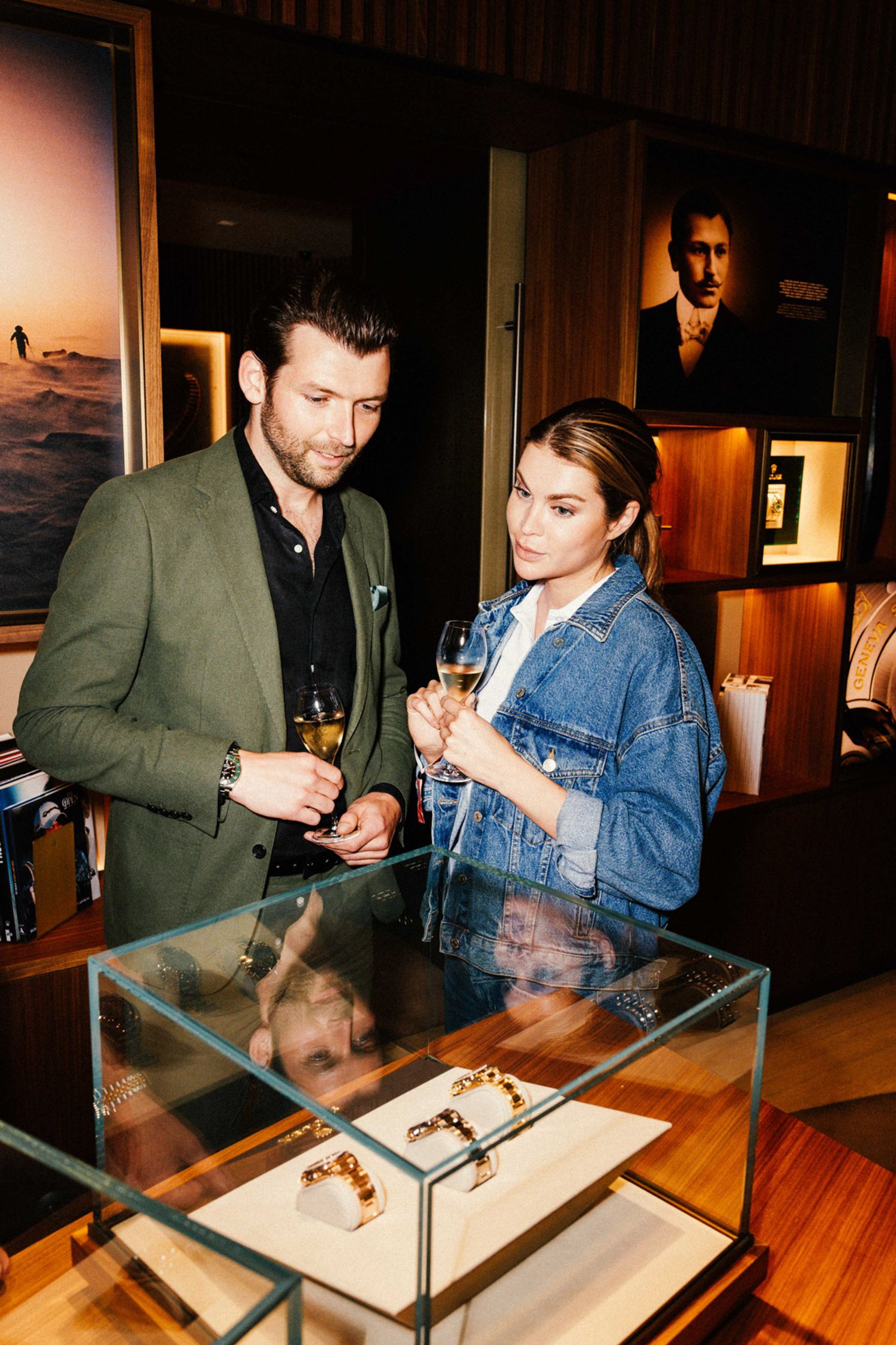Pärchen beim Rolex Boutique Event Berlin betrachtet goldene Rolex Uhren in einer Glasvitrine: Mann im olivgrünen Blazer und Frau in Jeansjacke halten Champagnergläser, Rolex Archivfotos im Hintergrund