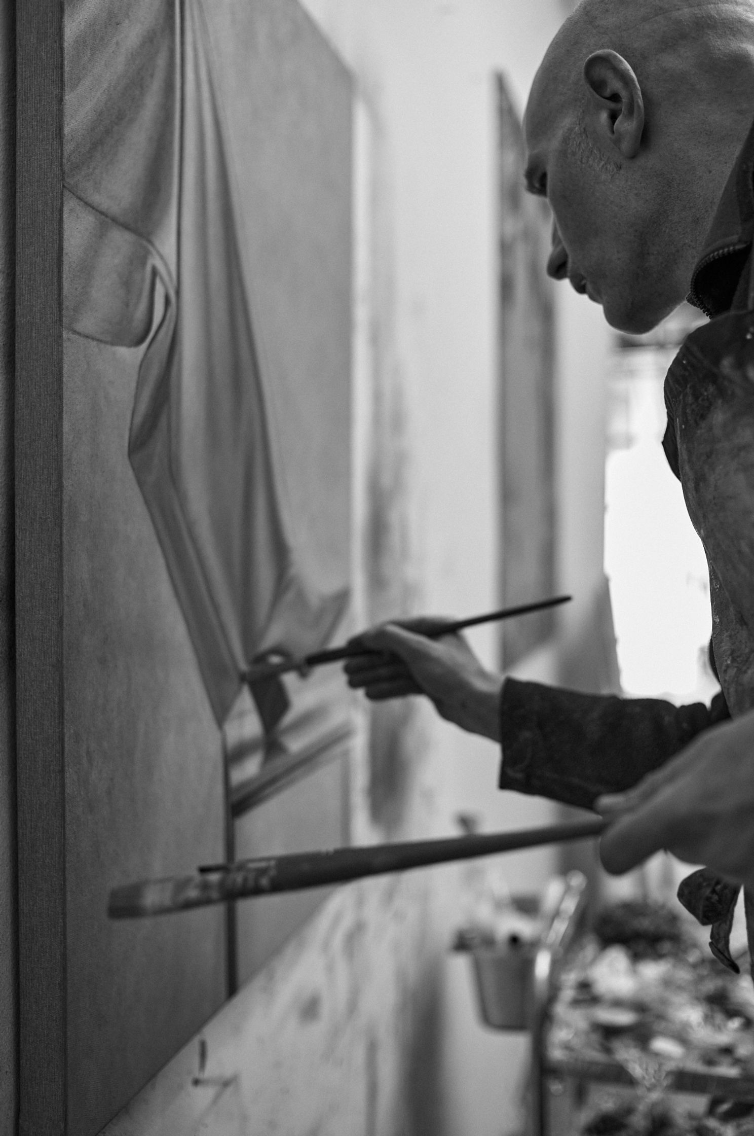 Schwarzweißaufnahme eines Künstlers in konzentrierter Arbeit. Ein Mann mit rasiertem Kopf steht seitlich vor einer Leinwand und malt mit einem feinen Pinsel an einer figurativen Darstellung eines menschlichen Körpers. In der anderen Hand hält er einen weiteren Pinsel. Der Fokus liegt auf seiner Handbewegung und der präzisen Berührung der Leinwand. Der Hintergrund ist unscharf, mit Andeutungen eines Ateliers und Malutensilien, wodurch eine ruhige, intime Arbeitsatmosphäre entsteht.