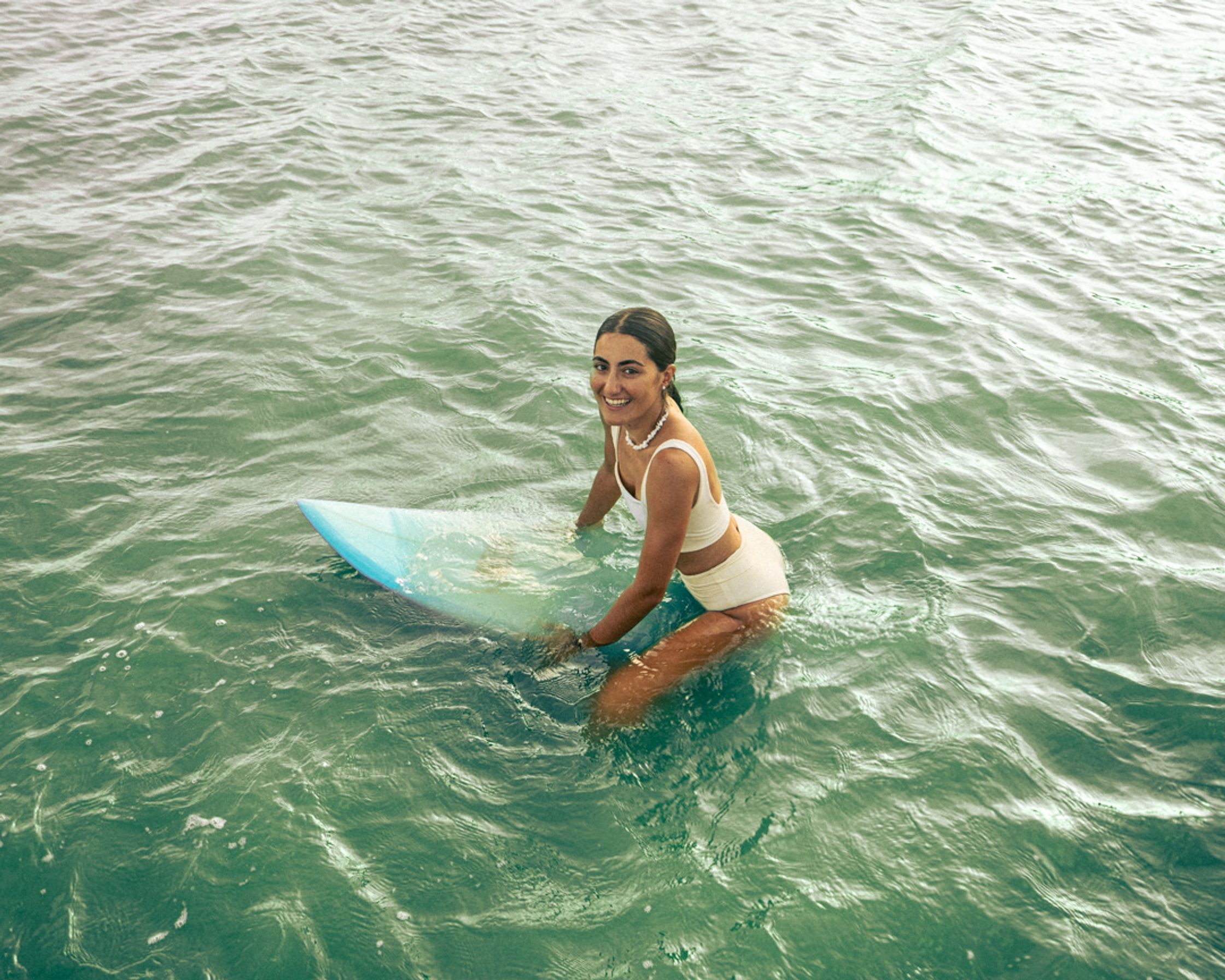 An authentic portrait of a woman sitting on her surfboard in the water at Kuta Lombok, looking directly and confidently at the camera.