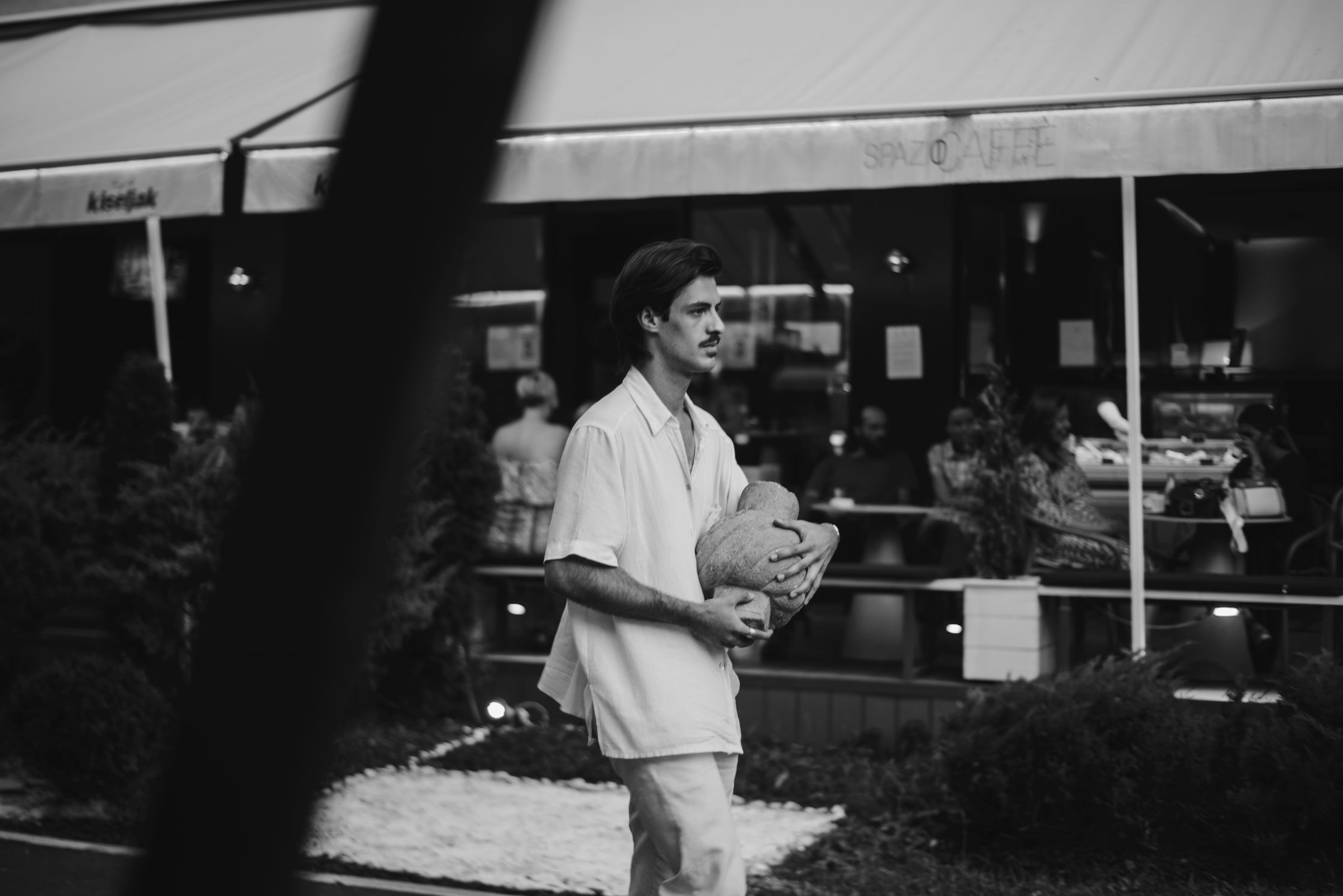 A black and white photo of a man in a white suit carrying a large gourd with storefronts and patrons in the background.