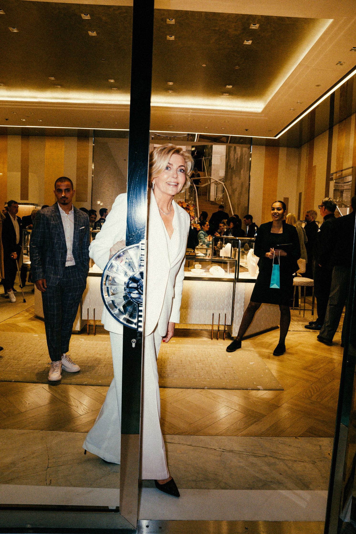 Elegante Dame im weißen Hosenanzug öffnet die Glastür der Tiffany & Co. Boutique Berlin – Gäste und Schmuckvitrinen im warmen Licht dahinter