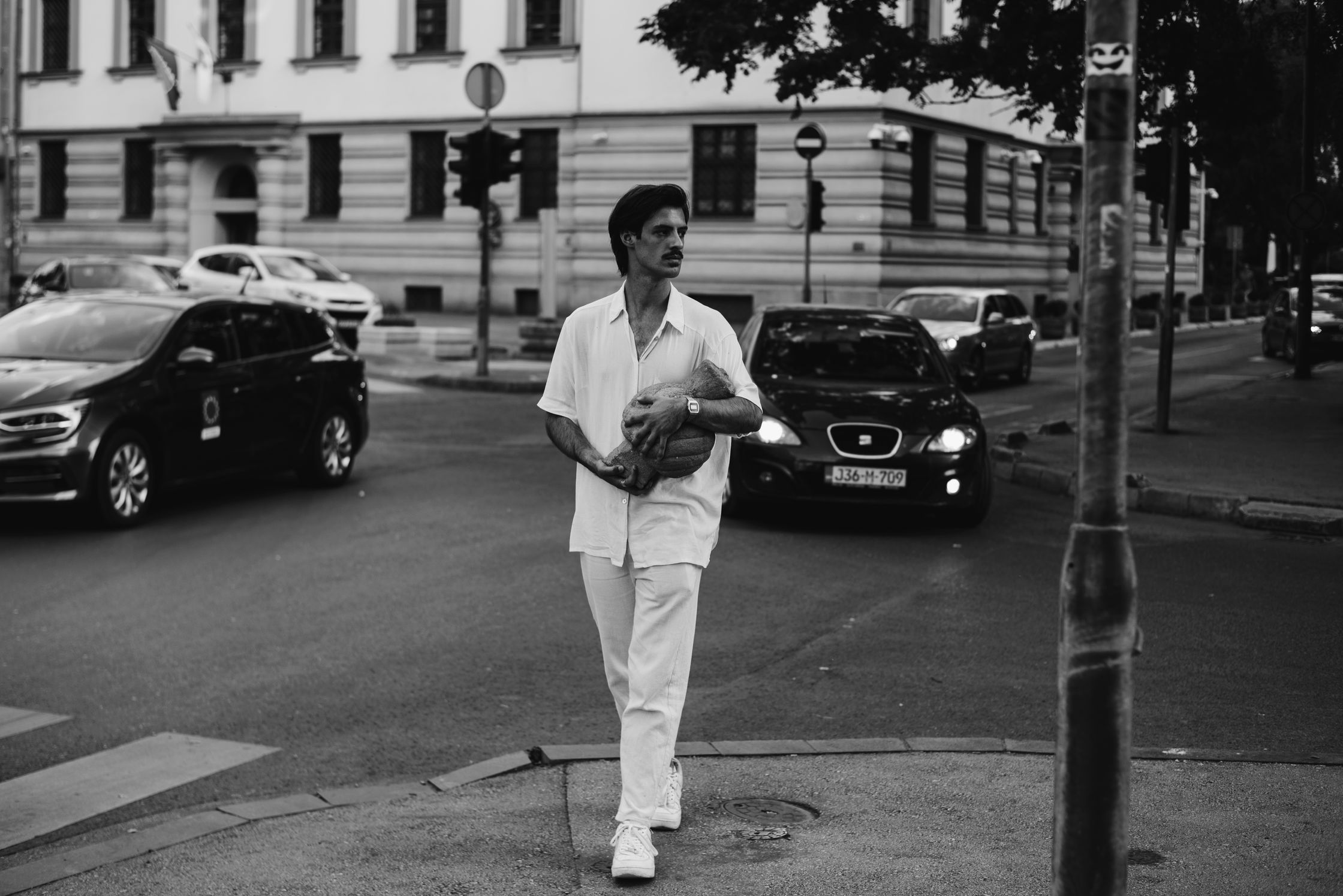 A man in a white outfit walks across a city street carrying a large, textured object, with cars and buildings in the background.