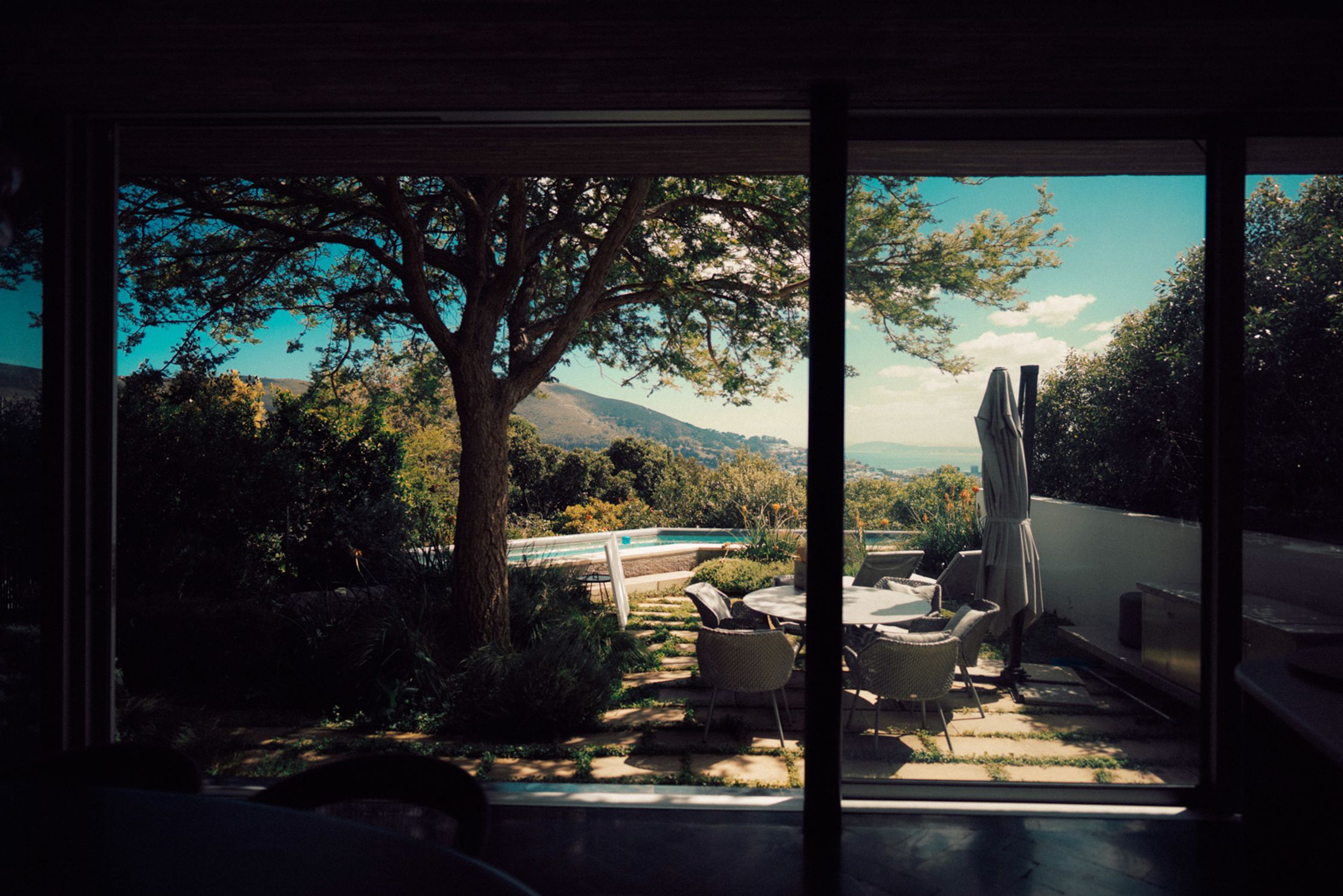 Blick durch Glasschiebetüren einer modernen Kapstädter Villa auf Terrasse mit Loungemöbeln, Pool, großem Baum und Bergpanorama im Hintergrund, Lifestyle-Shoot Paul Schrader Cape Town 2024