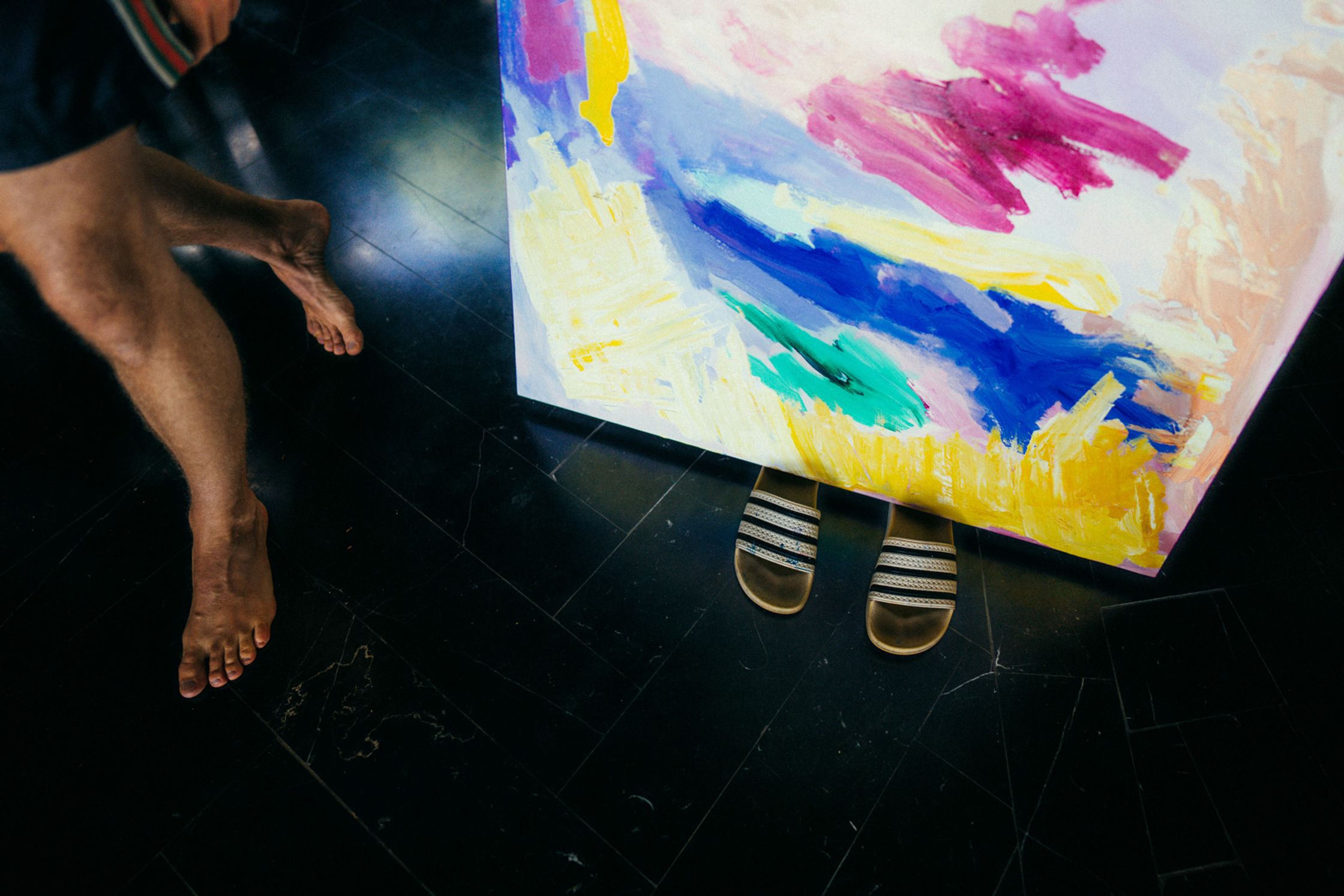 Detail-Aufnahme von Paul Schraders Füßen neben einem großen bunten abstrakten Gemälde auf dunklem Boden, leuchtende Farben Lila, Gelb und Blau, intimes Künstlerportrait Kapstadt 2024