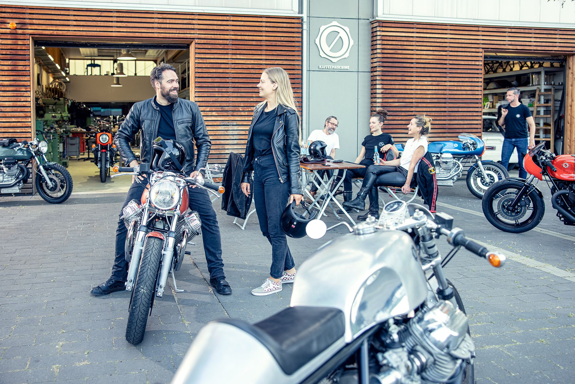 A group of motorcyclists relaxing outdoors in front of a garage-style cafe with several custom motorcycles parked nearby.