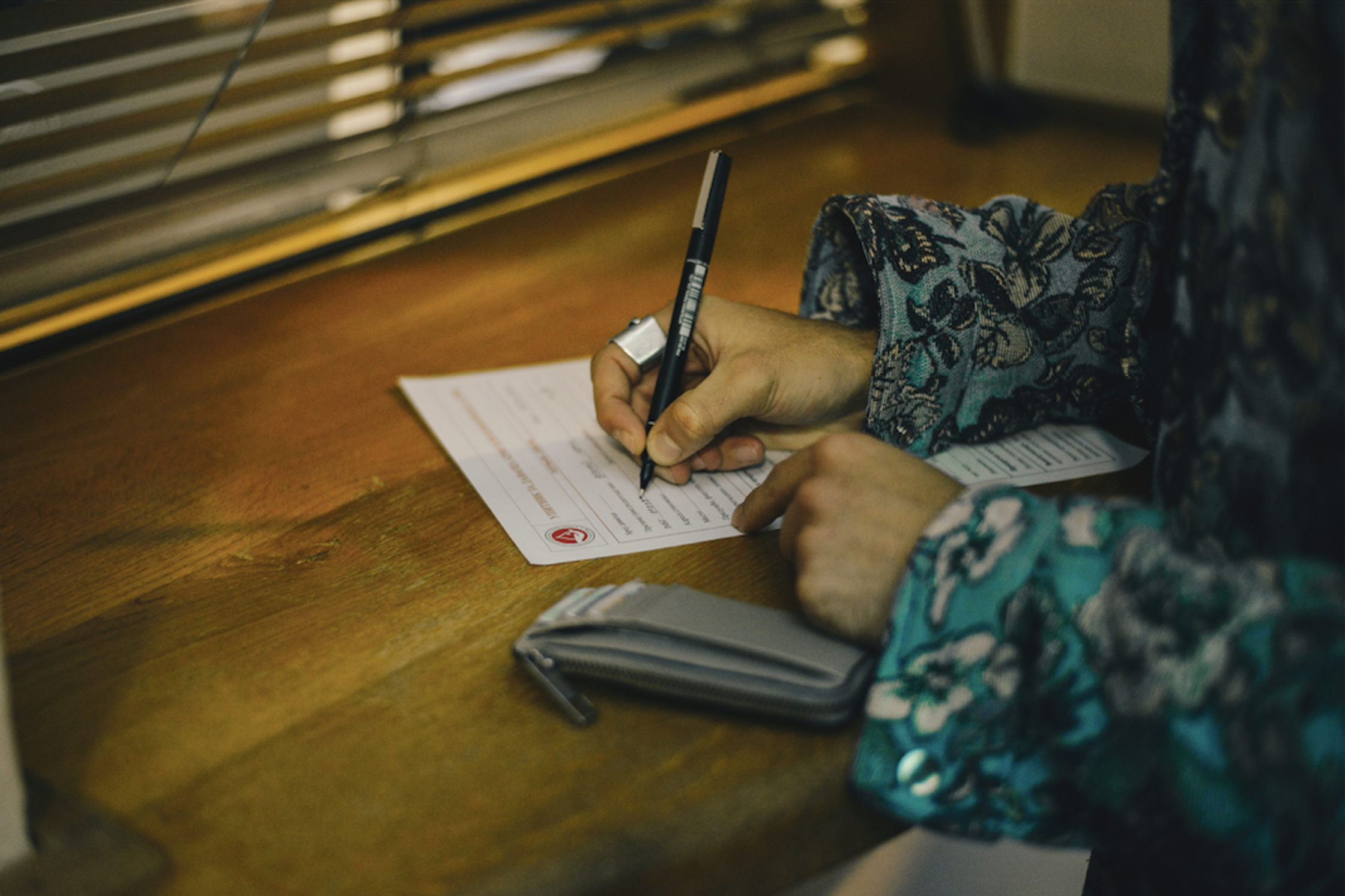A person with a patterned sleeve writes on a document with a pen on a wooden table.