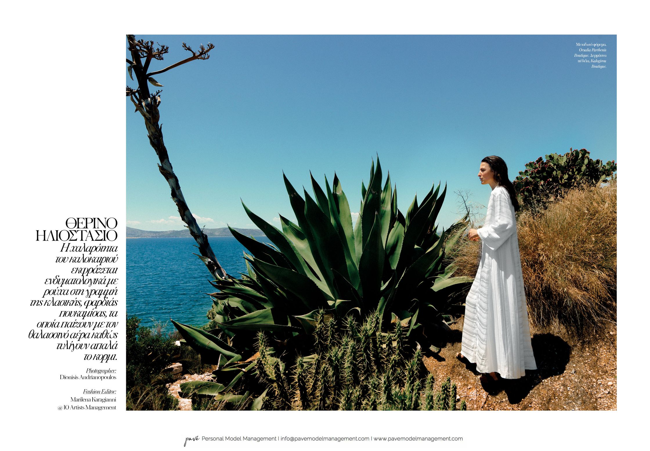 A woman in a long white dress stands on a rocky path overlooking the blue sea, surrounded by large succulents and dry grasses under a clear sky.