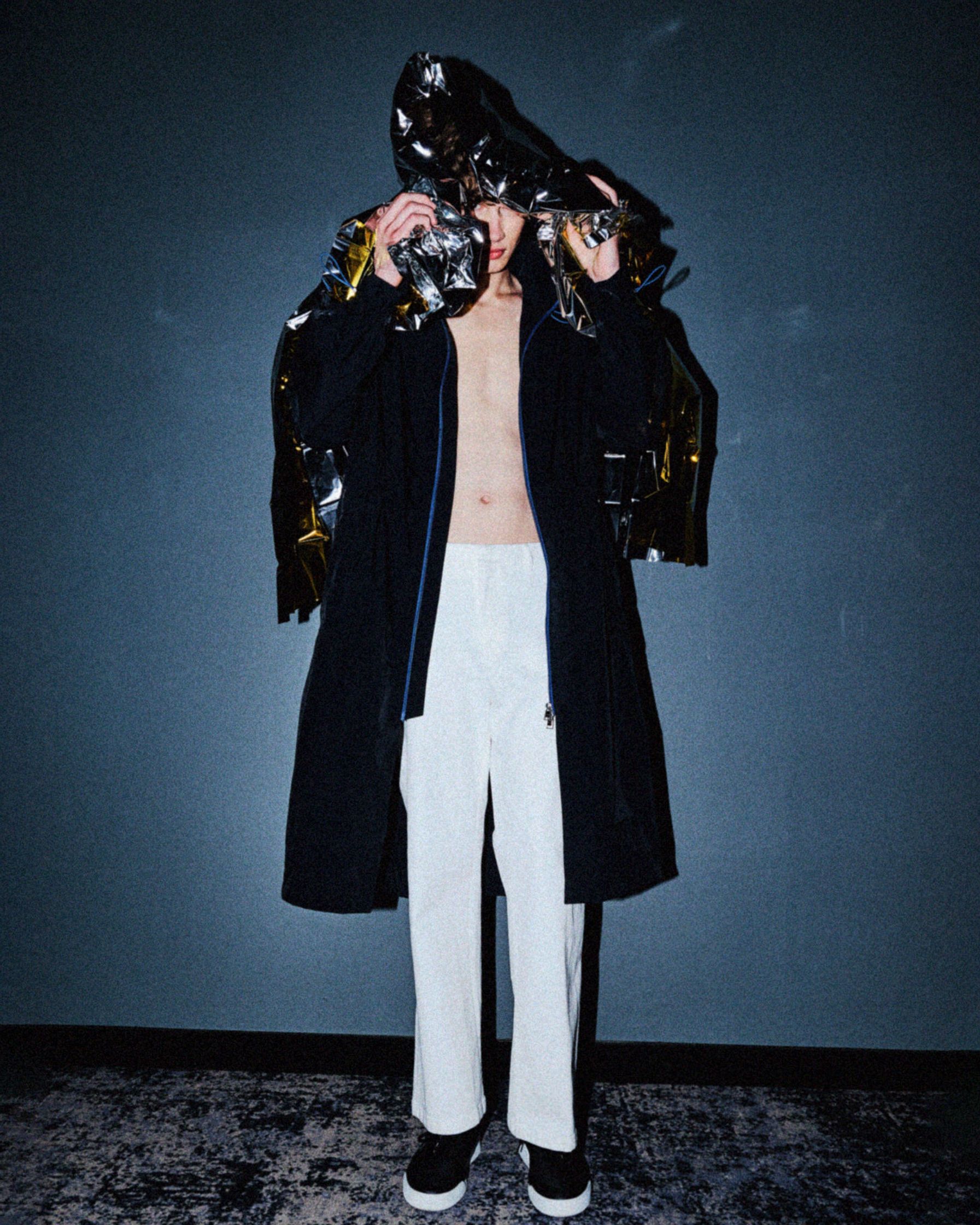 a man in a black coat and white pants standing in front of a black wall