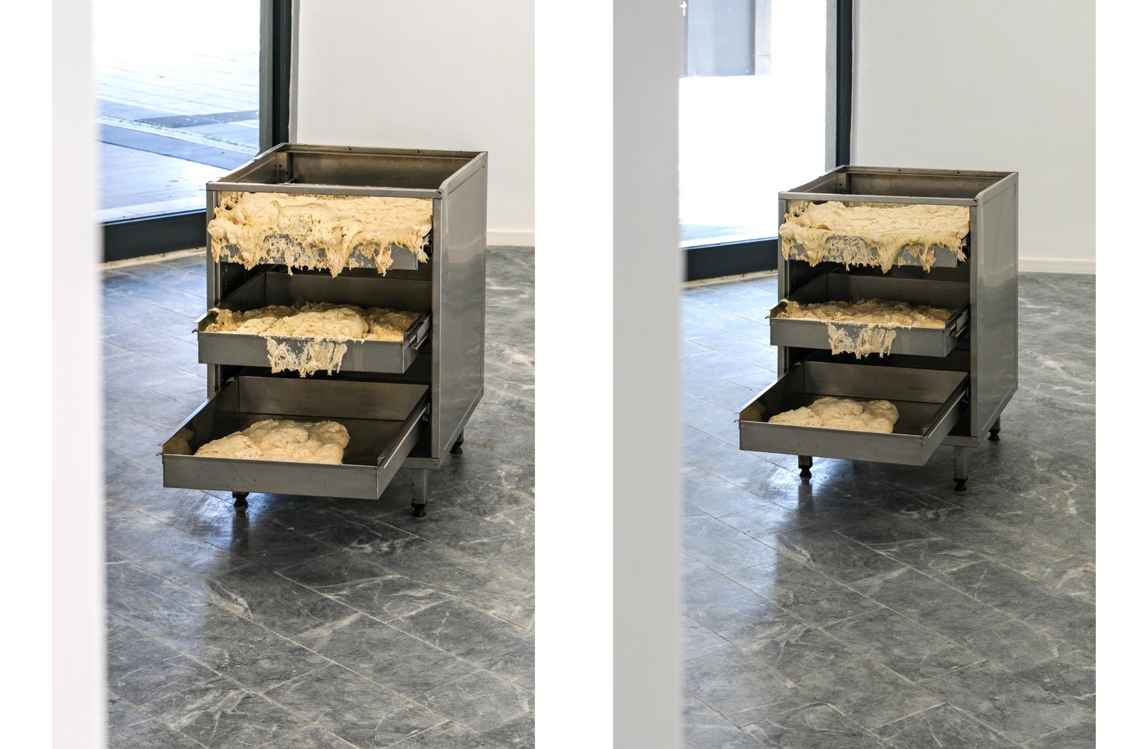 Fluffy white dough overflows in three metal trays, set within a stainless steel cabinet on a tiled floor.