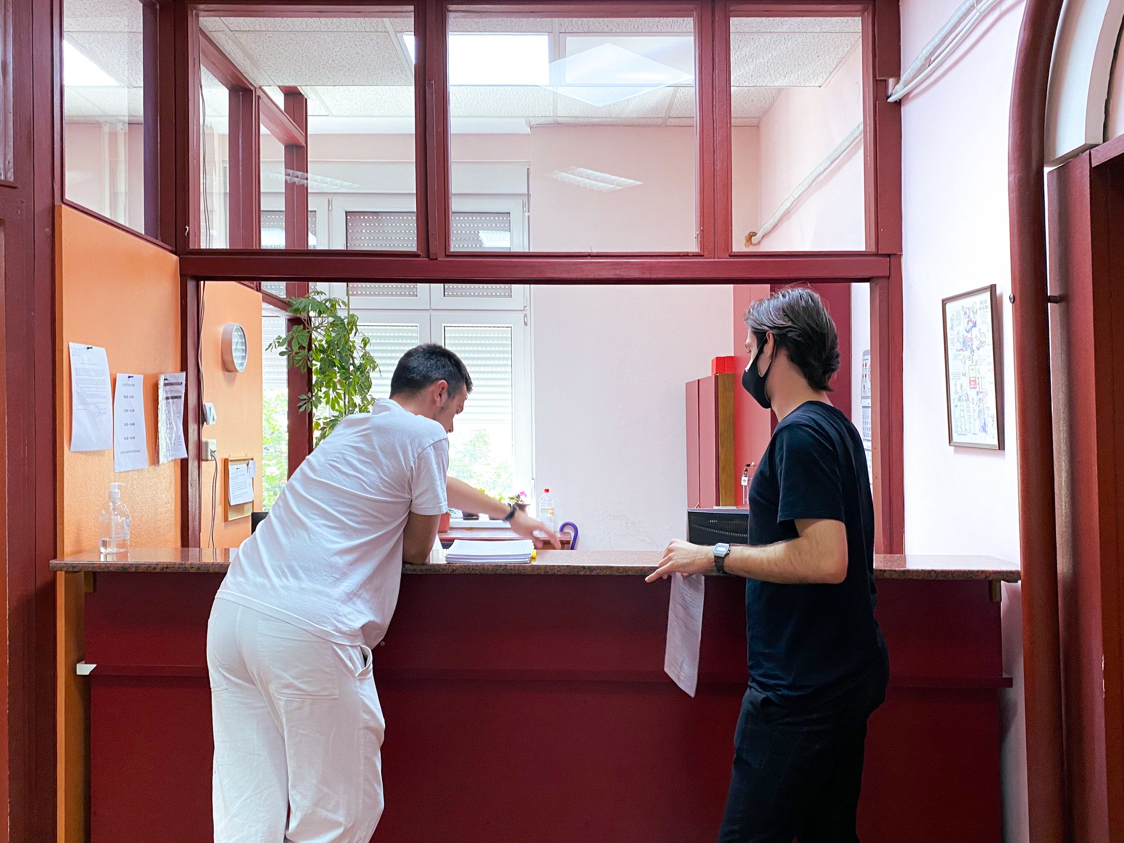 A man in a black mask speaking to a man in a white uniform at a reception desk.