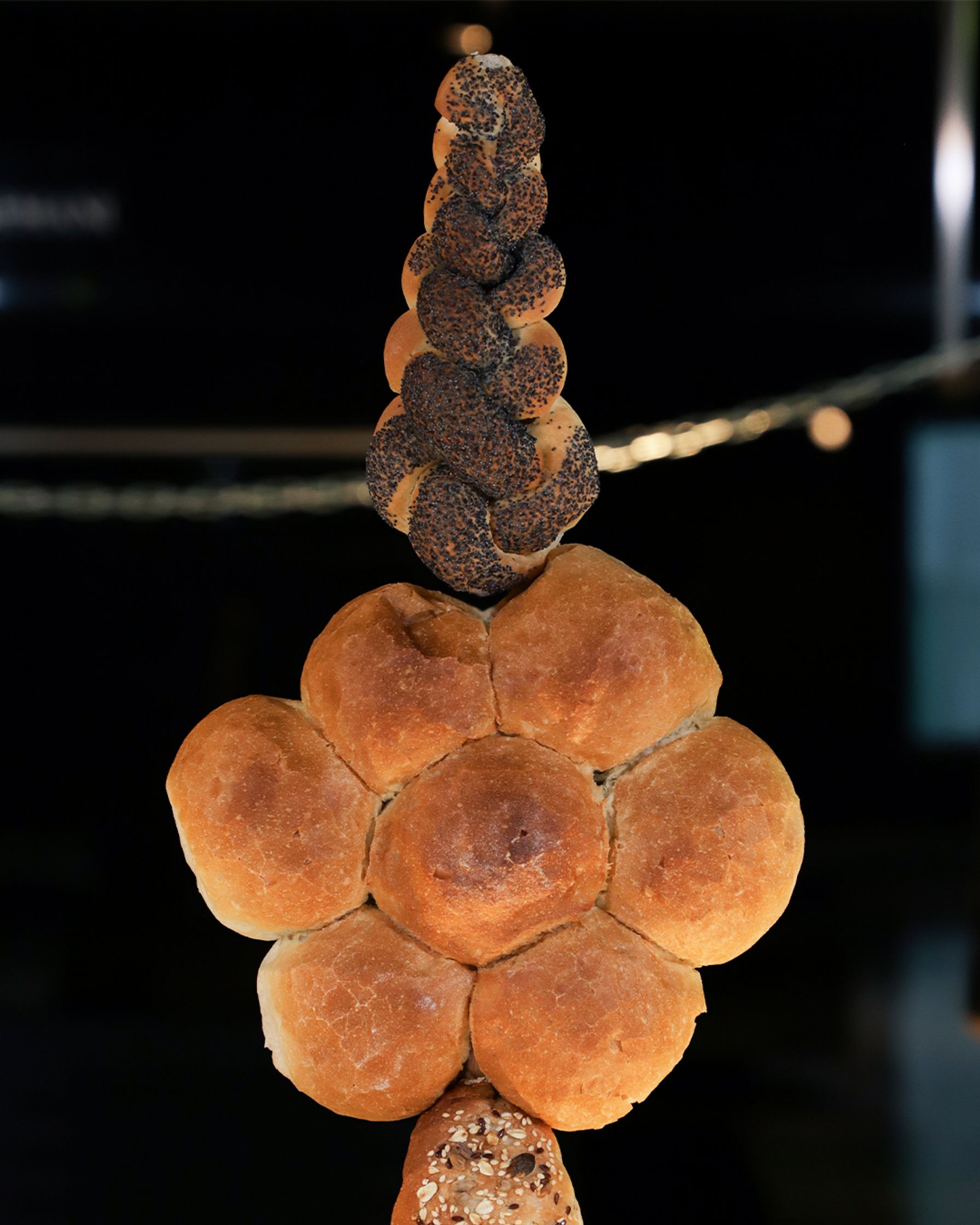 A decorative display of bread loaves, with a long braided poppy seed loaf at the top, round, golden-brown rolls in the middle, and multi-grain bread at the bottom, against a dark, blurry background with subtle lights.