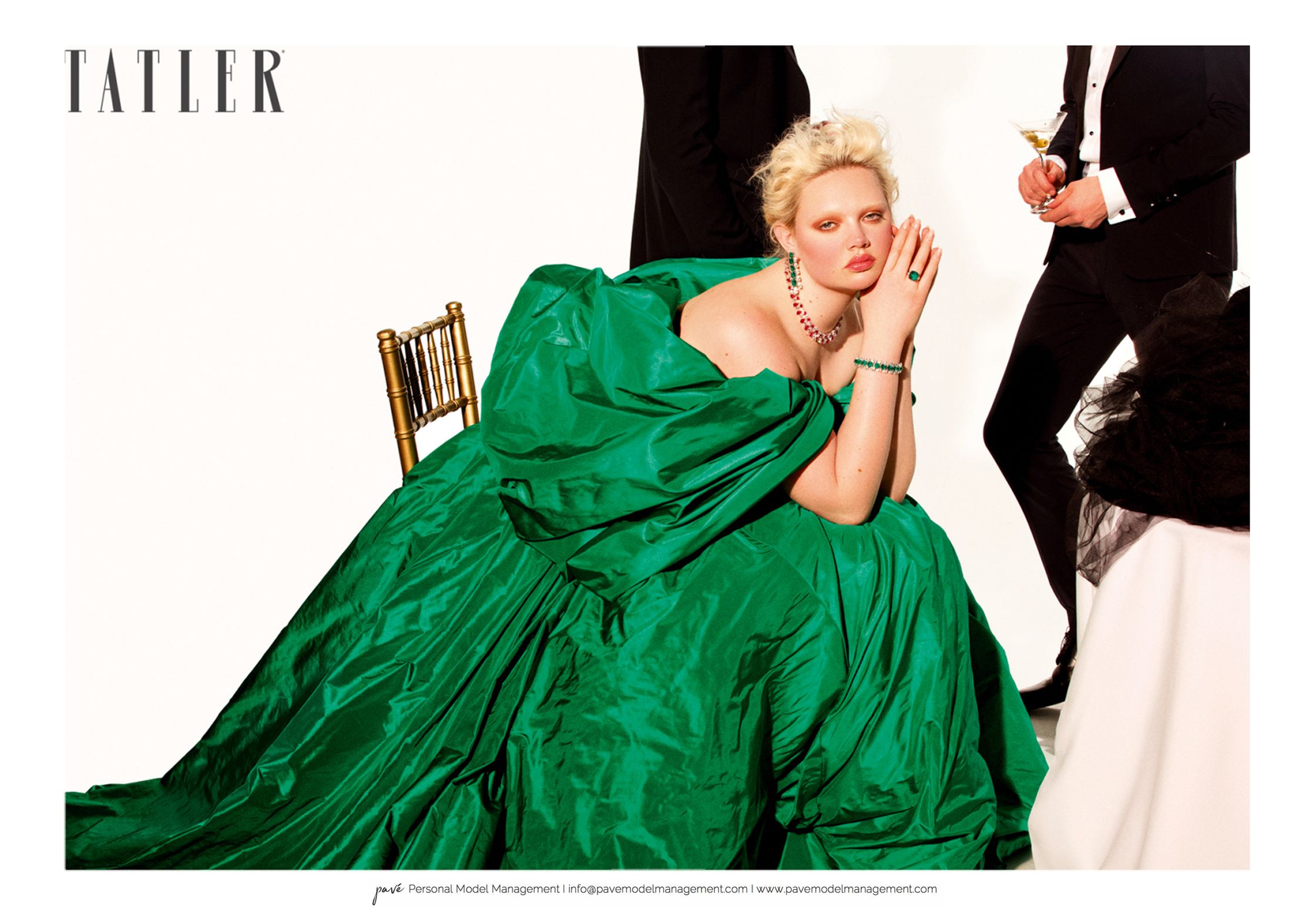 A fair-skinned person in an emerald off-the-shoulder gown and jewelry poses at the center while two people in black suits stand behind them with the Tatler logo in the top left.