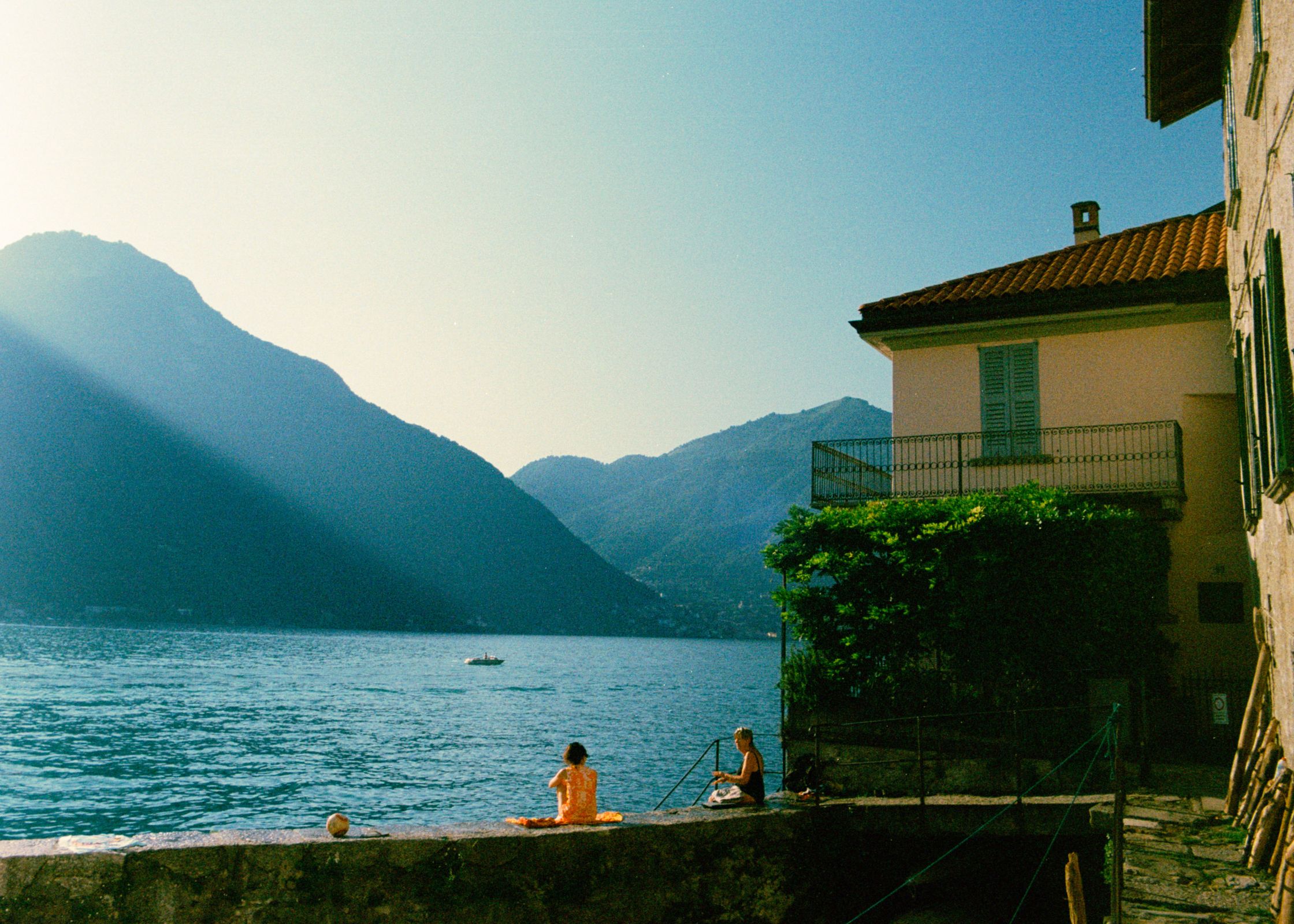 Candid photography of two friends sharing a moment of quiet contemplation on a stone dyke. A peaceful scene where women face the majestic mountains and the shimmering water of Lake Como.