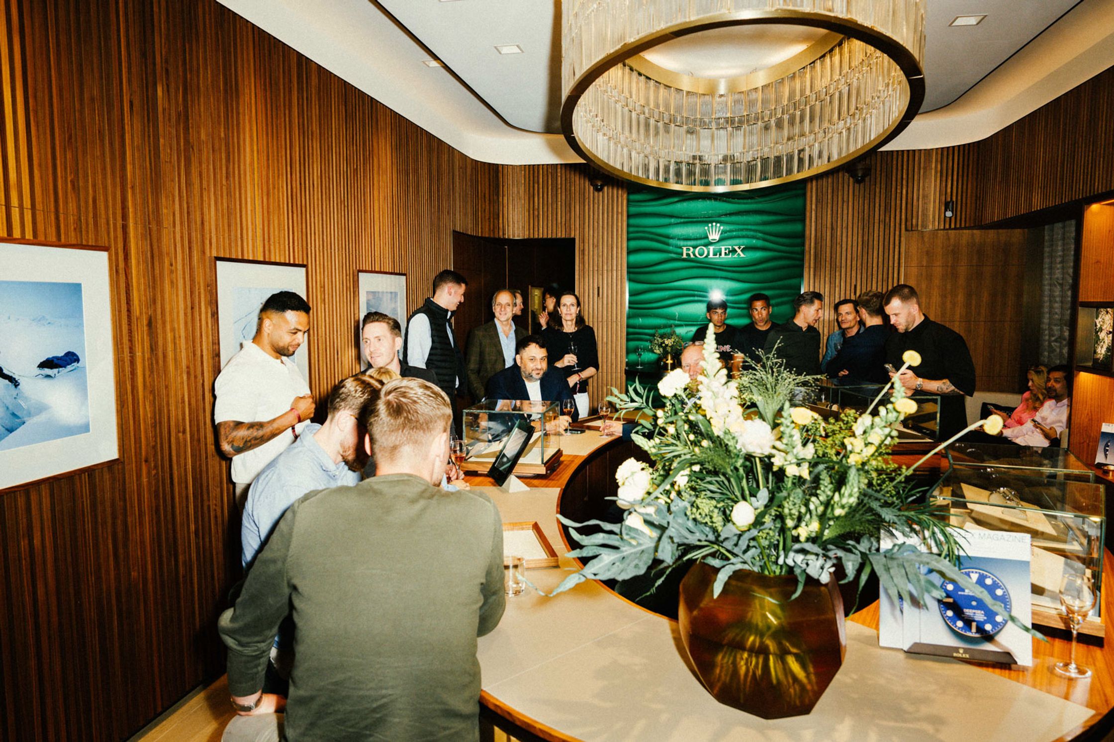 Exklusiver Abend-Event in der Rolex Boutique Berlin: Gut gekleidete Gäste um ovalen Uhren-Tresen mit Blumenarrangement, Rolex-Logo auf grüner Wand und elegantem Kristallleuchter