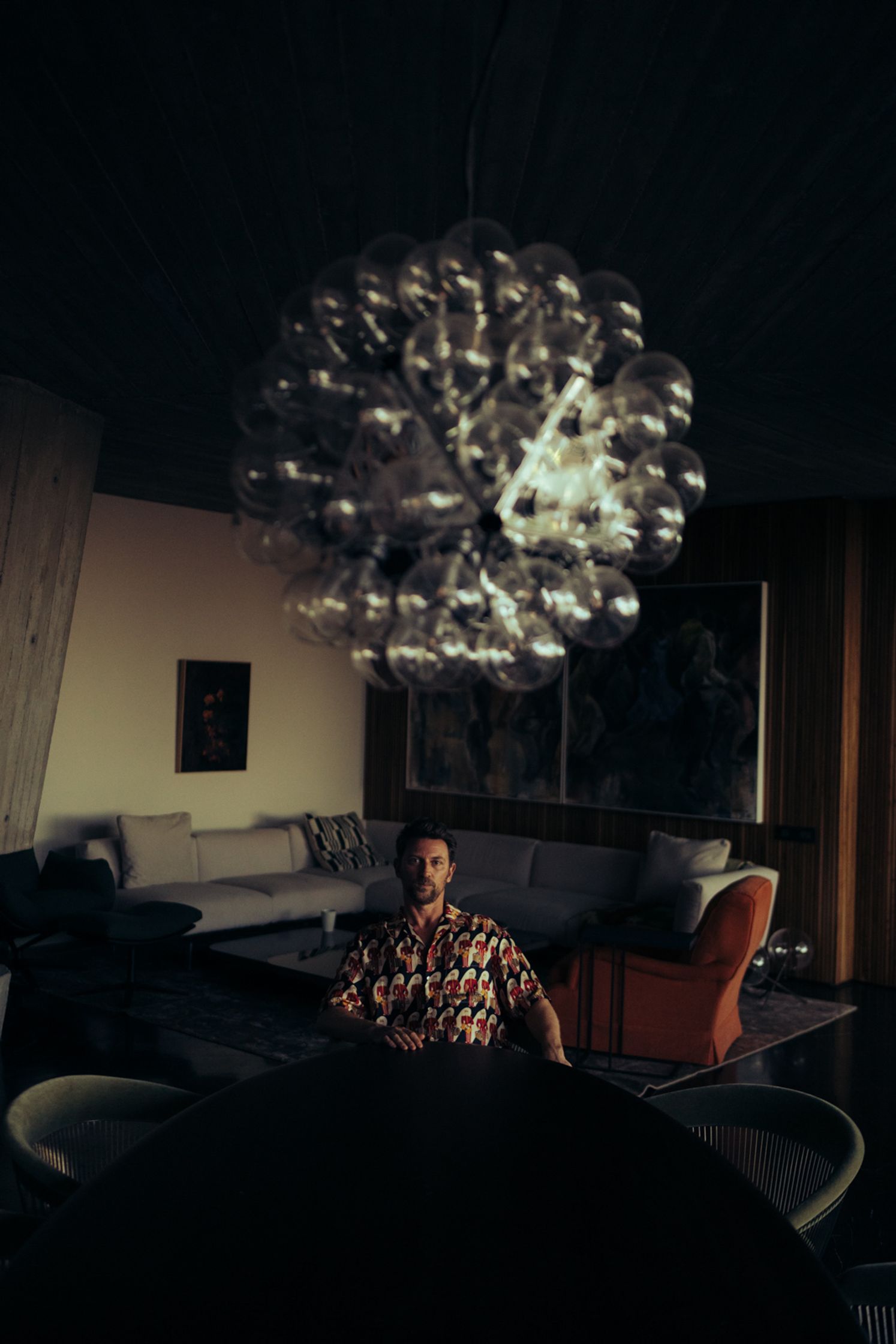 Künstler Paul Schrader sitzt unter einem spektakulären kugelrunden Designer-Kronleuchter in dunklem elegantem Interieur mit Sofa, orangem Sessel und Holzvertäfelung, Kapstadt 2024