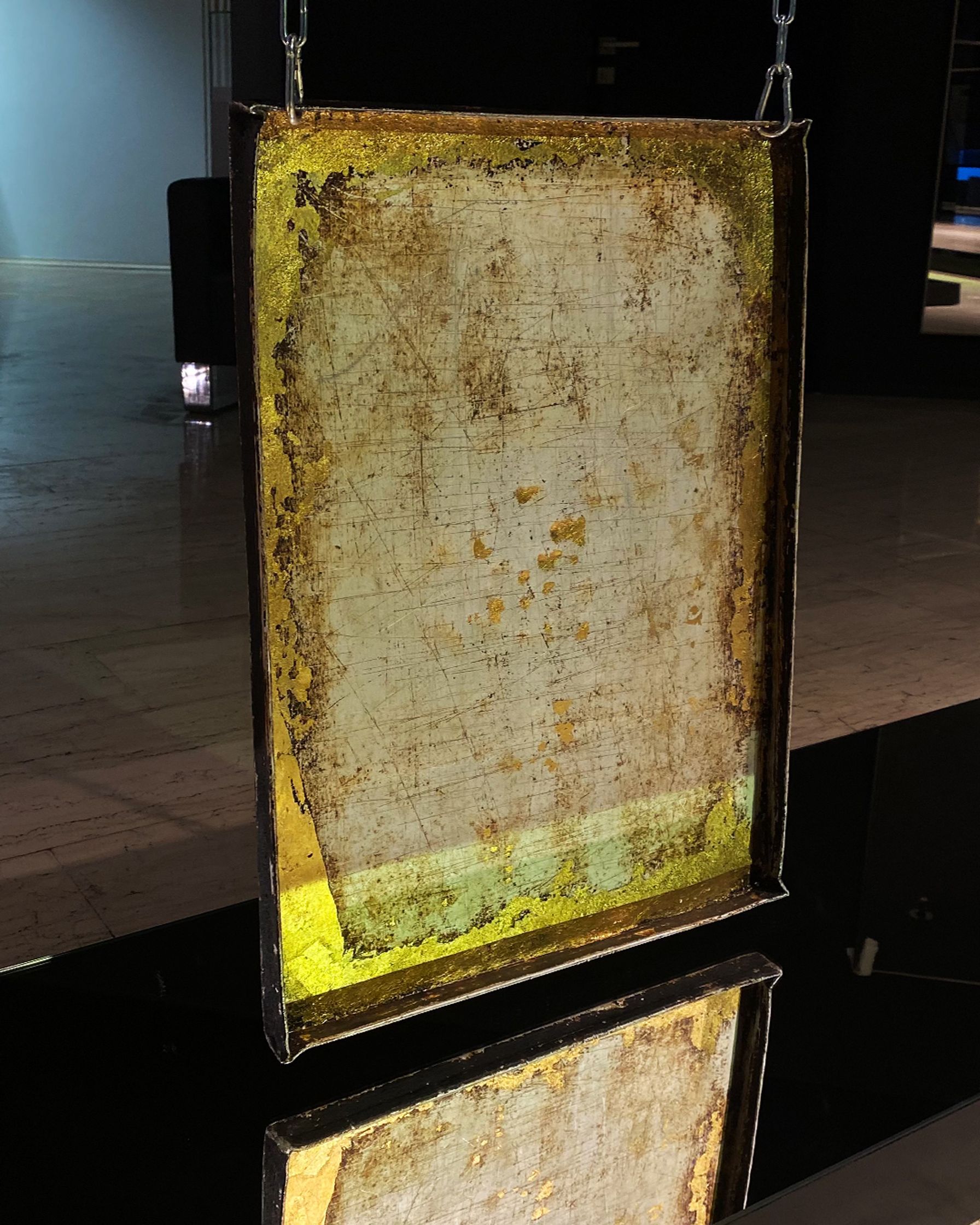 A rectangular piece of art, textured with scratches and a distressed, peeling gold and green border, hangs by a chain with a reflection beneath it.