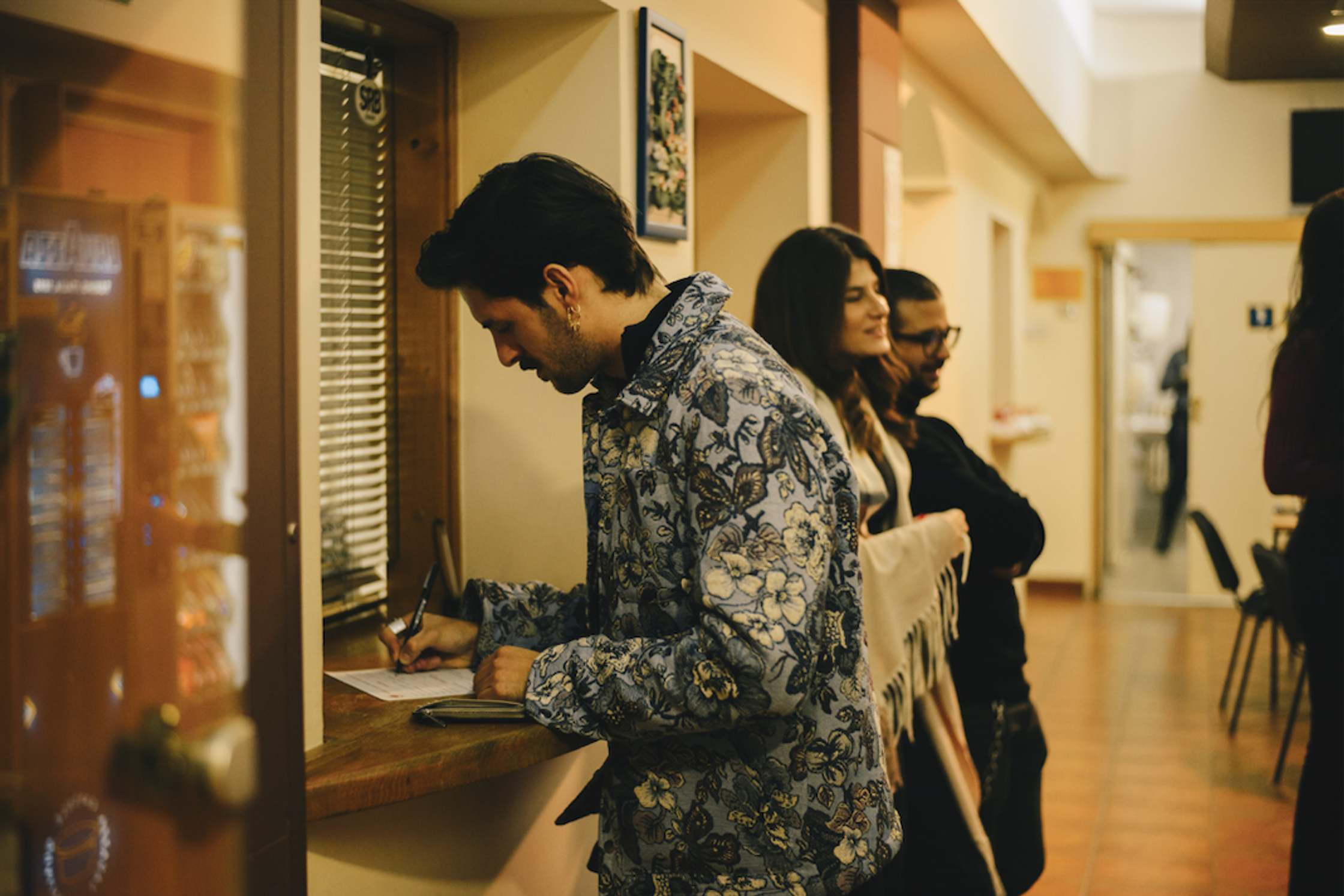 A man in a floral jacket signs a document at a counter as people wait in line behind him.
