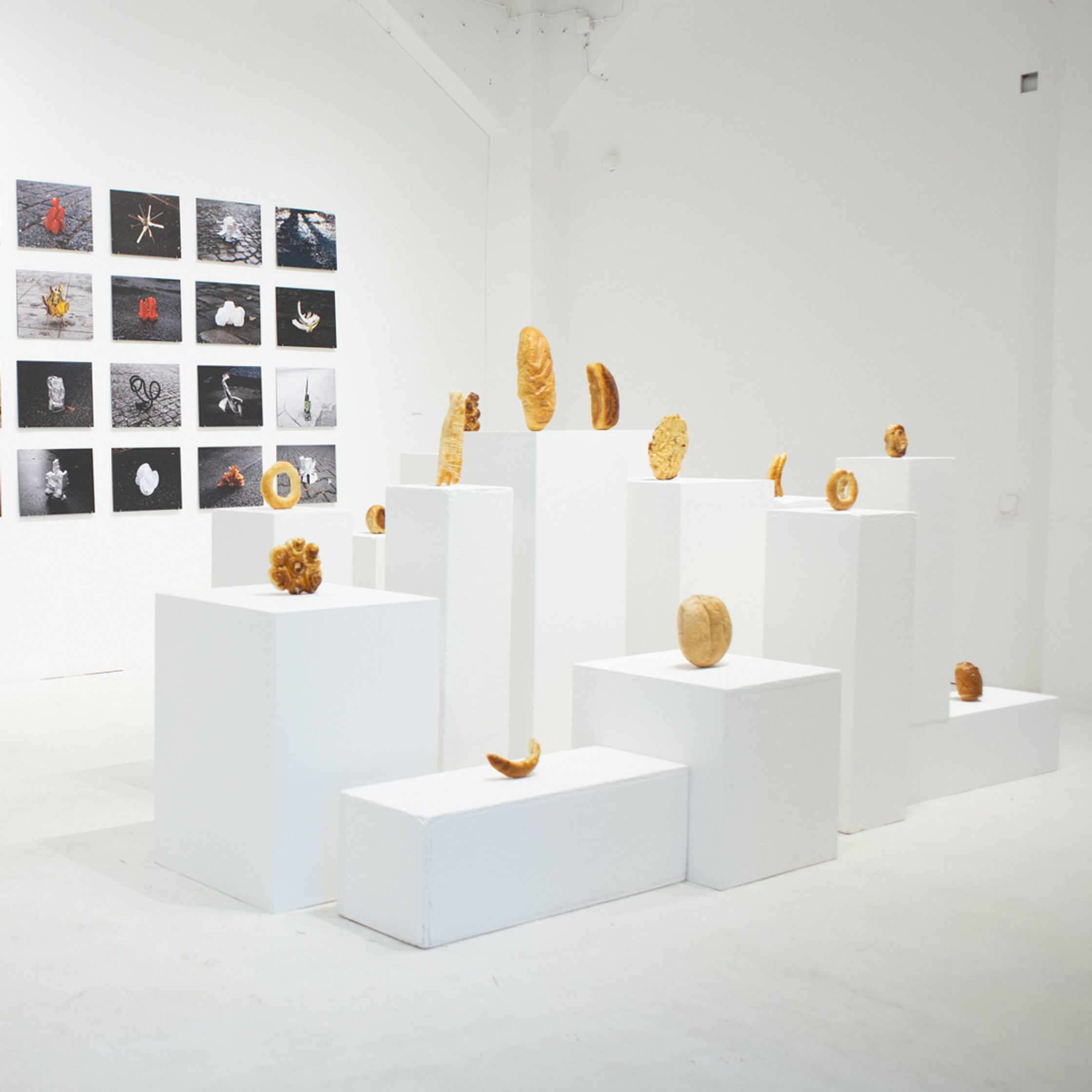 An art gallery with numerous bread and pastry-like sculptures on white pedestals and an arrangement of small, grid-like photographs.